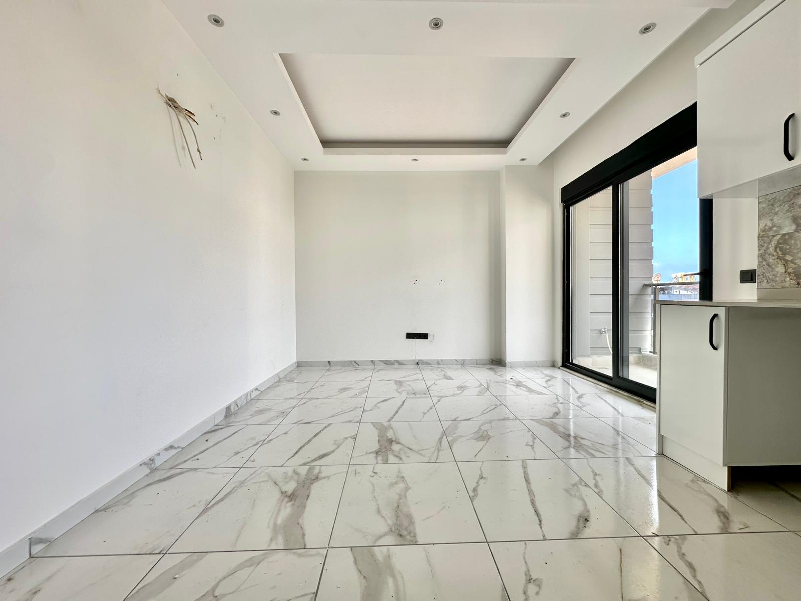Satılık konutun mutfaklı salonunda boş oturma alanı, balkon kapısı ve sağ duvarda dolaplar