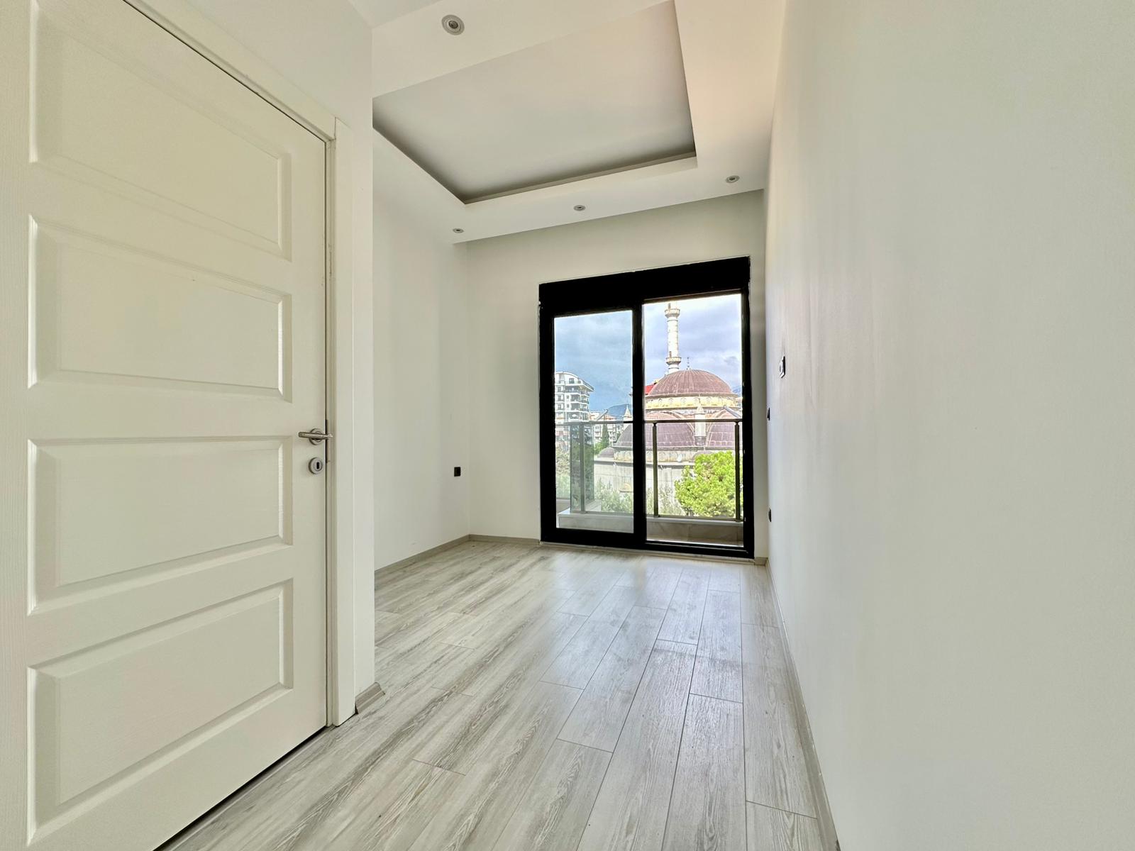 Alanya’daki dar yatak odasında siyah çerçeveli balkon kapısı ve pencereden kubbe ile minare görünümü
