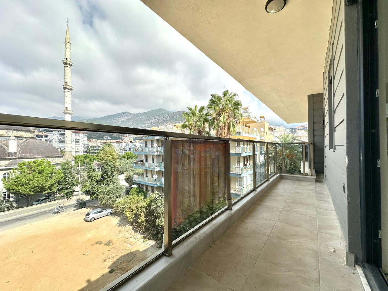 Satıştaki yapının uzun kapalı balkonunda cam korkulukların ardından dağlar ve minare görünümü