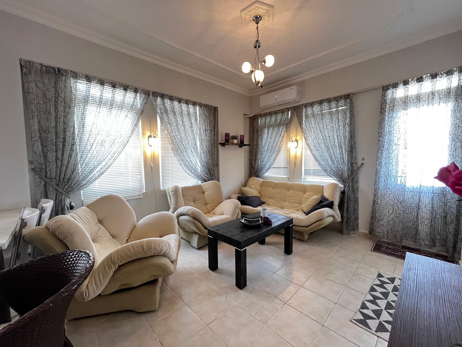 Alanya’daki dairenin salonunda krem kanepe, iki koltuk ve desenli perdeli üç pencere