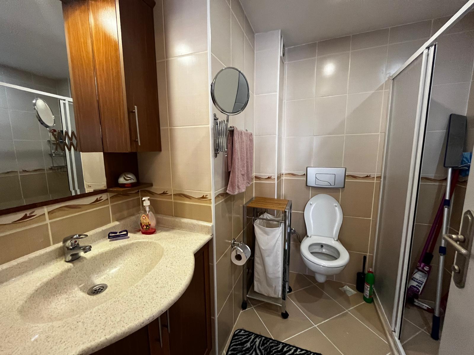 Alanya’daki dairenin banyosunda cam kapılı duş kabini, lavabolu dolap ve çapraz döşenmiş karo