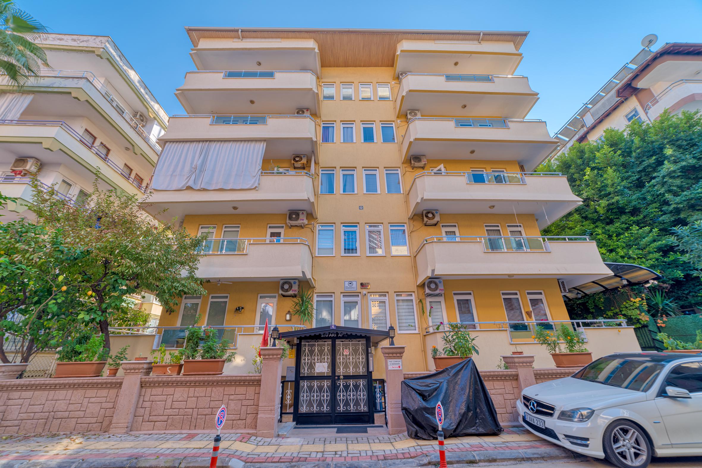 Alanya’daki dairenin sarı beş katlı cephesi, cam korkuluklu geniş balkonlar ve siyah giriş kapısıyla