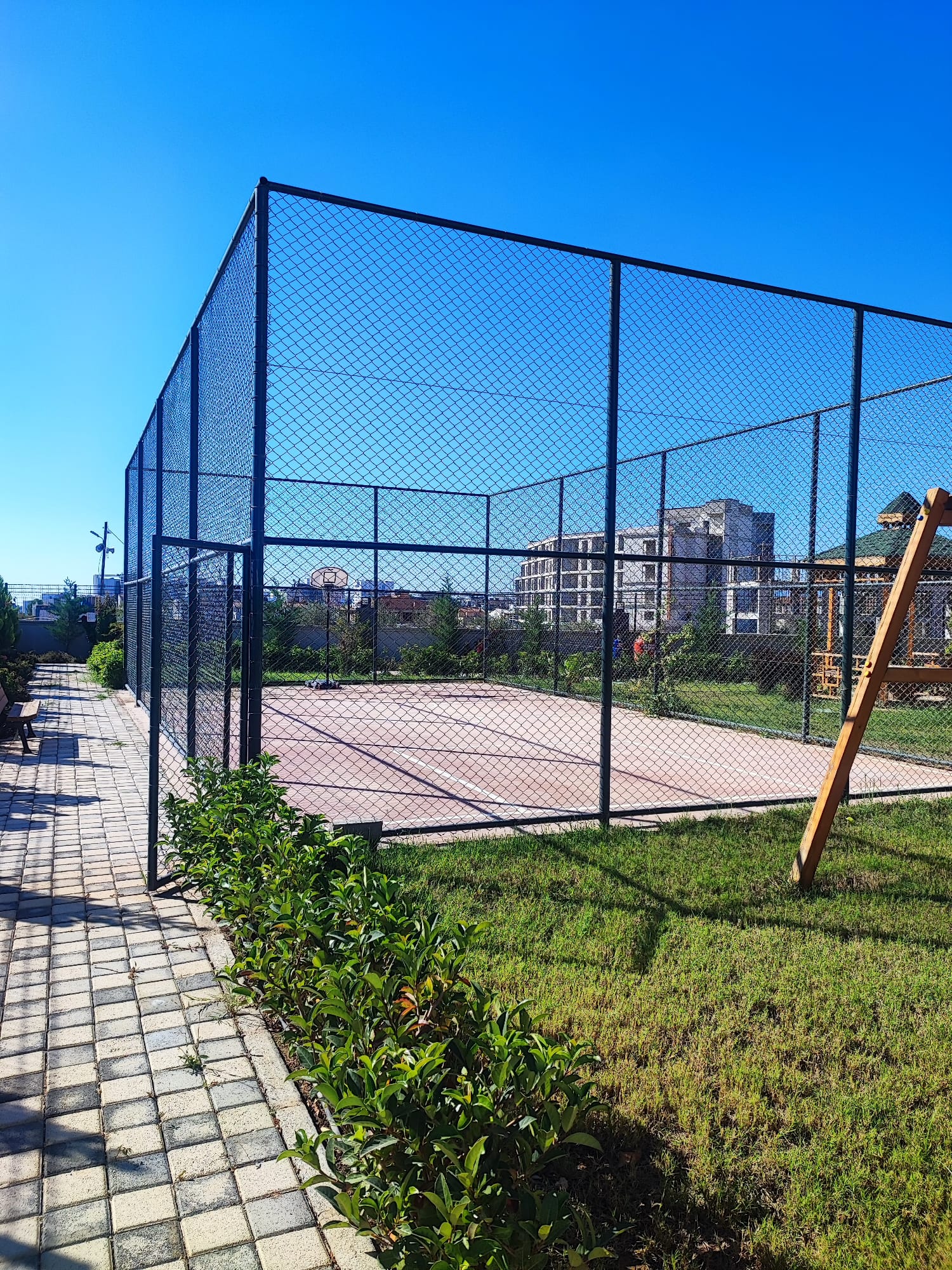 Fenced sports court with a basketball hoop and pink surface on landscaped grounds in Antalya