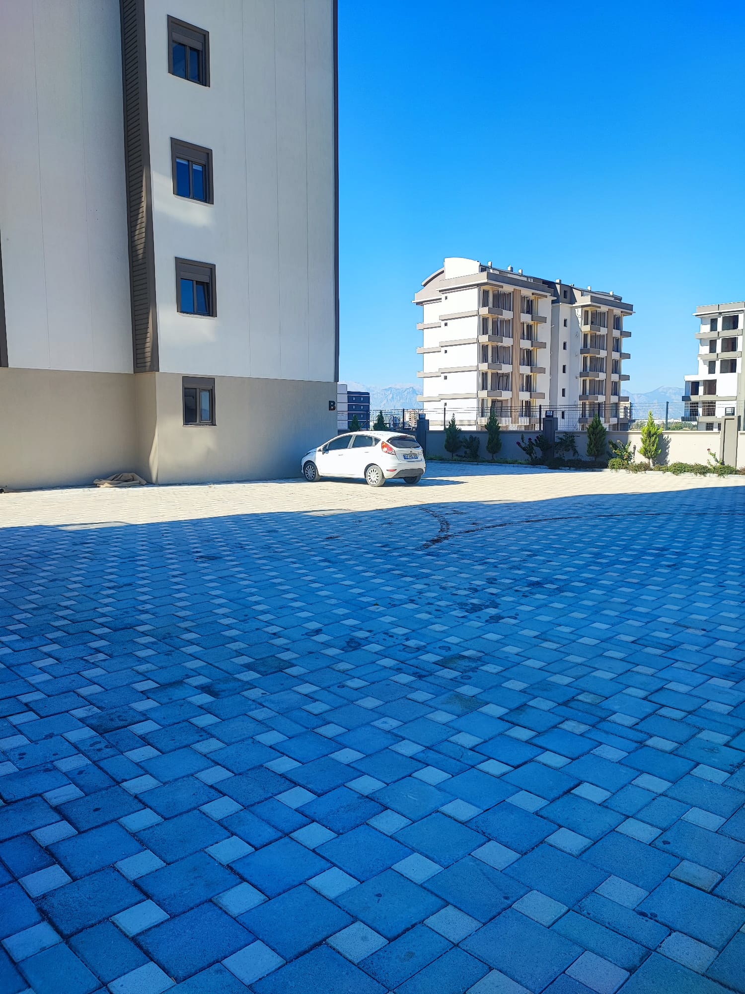 Paved parking area with a white car near residential buildings with 1+1 flats in Altyntaş