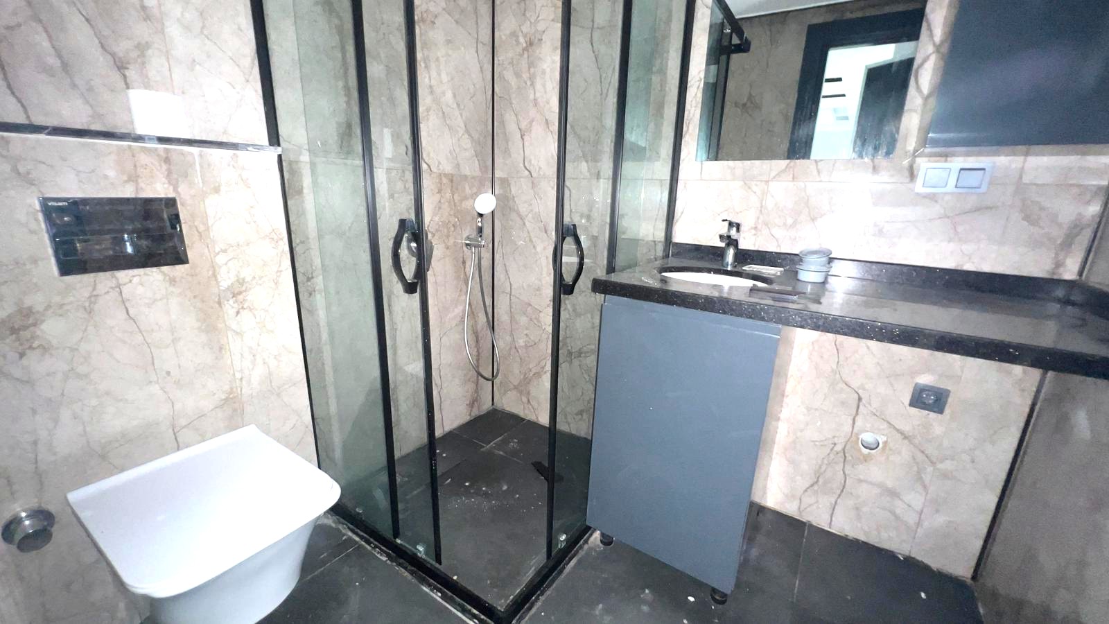 Alanya’daki banyoda cam köşe duş kabini, gri lavabolu dolap ve mermer desenli duvarlar