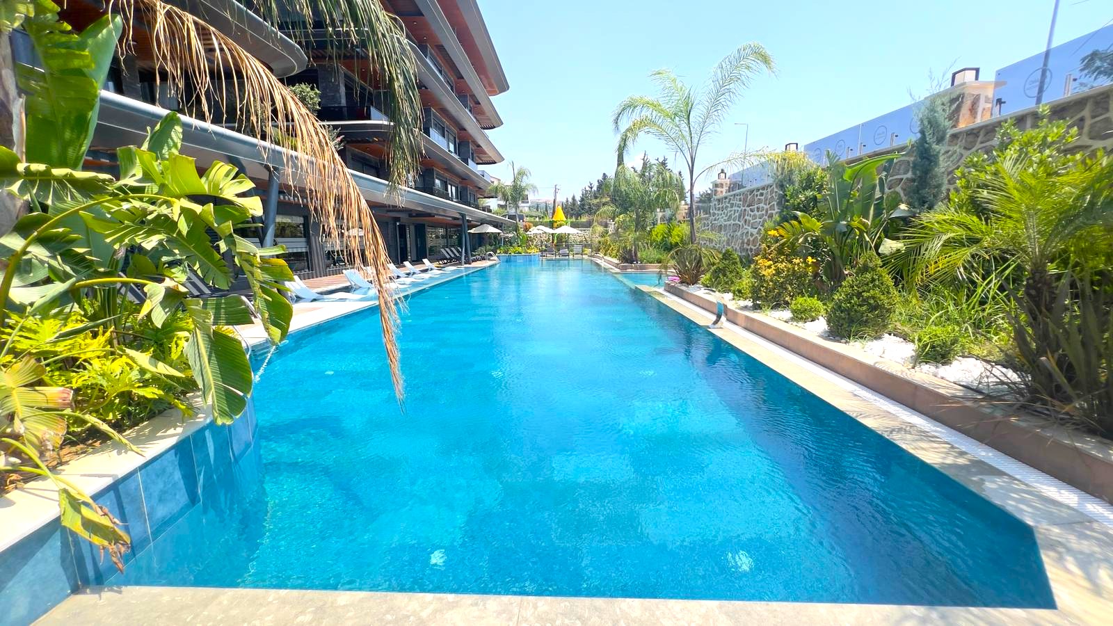 Alanya’da rezidansın açık havuzunda palmiyeler, taş çevre duvarı ve su boyunca uzanan açık renkli döşeme