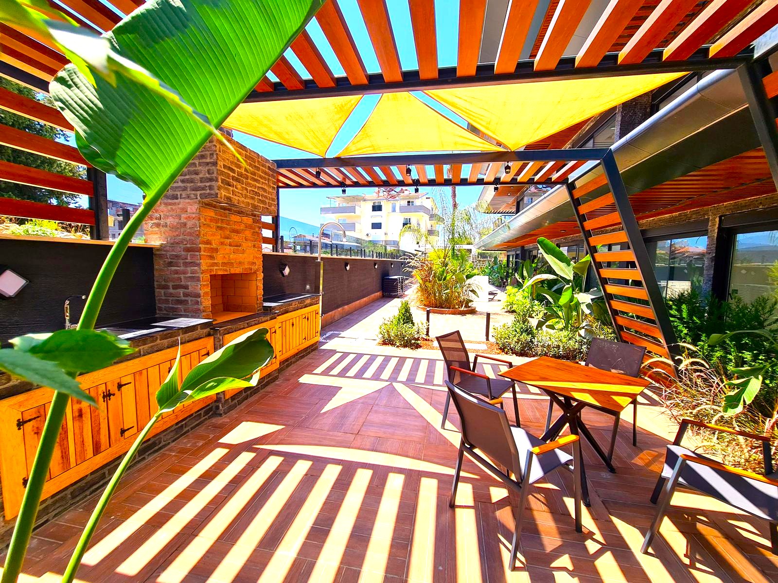 Alanya’da satılık dairelerin olduğu sitede sarı tentili pergola, tuğla barbekü ve ahşap yemek masası