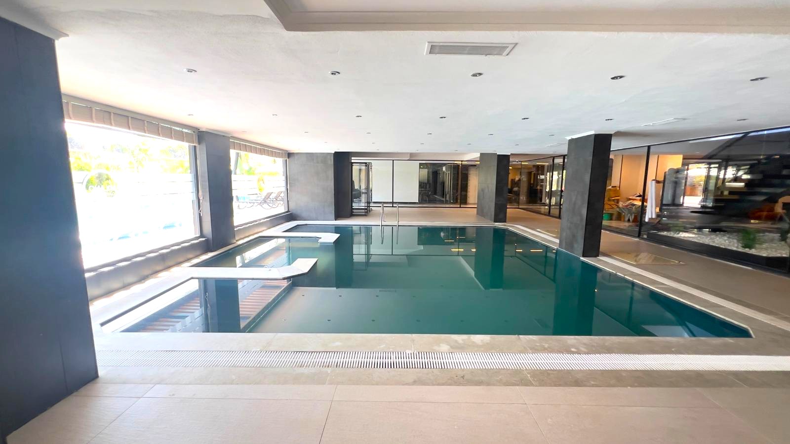 Alanya’daki konut kompleksinin kapalı havuzunda turkuaz su, basamaklar ve havuz boyunca büyük pencereler