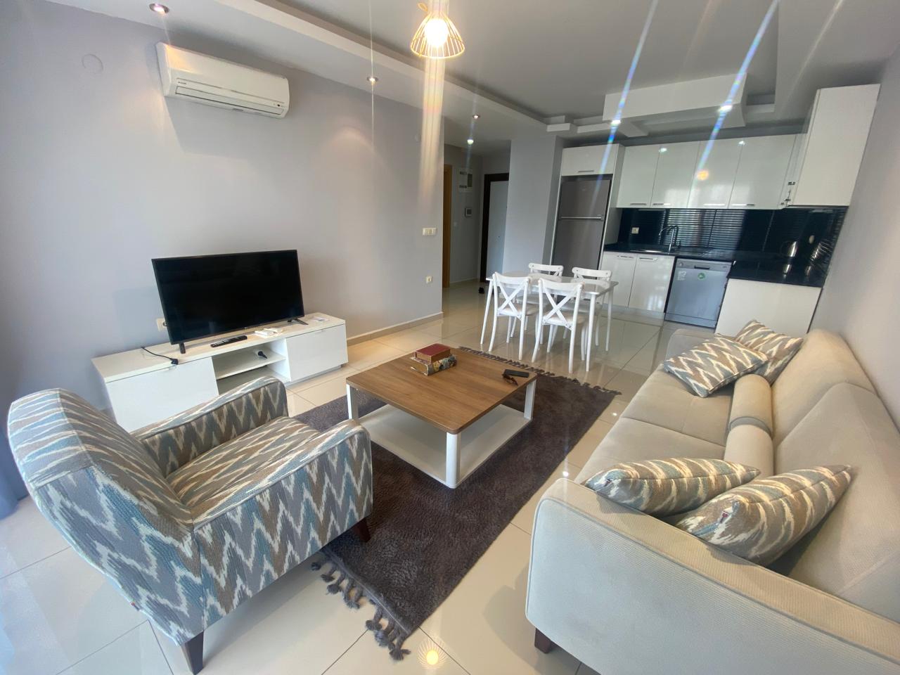 Kiralık dairenin mutfak-salonunda açık renk köşe koltuk, beyaz mutfak ve yemek masası bulunuyor