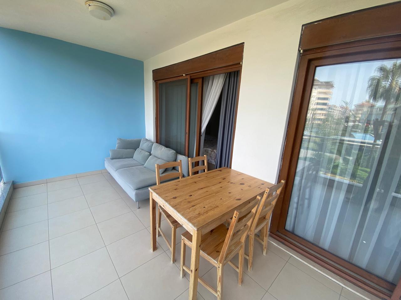 Ahşap masa ve gri kanepeli balkon Türkiye’de kiralama yapılan konutta şehir binalarına bakıyor