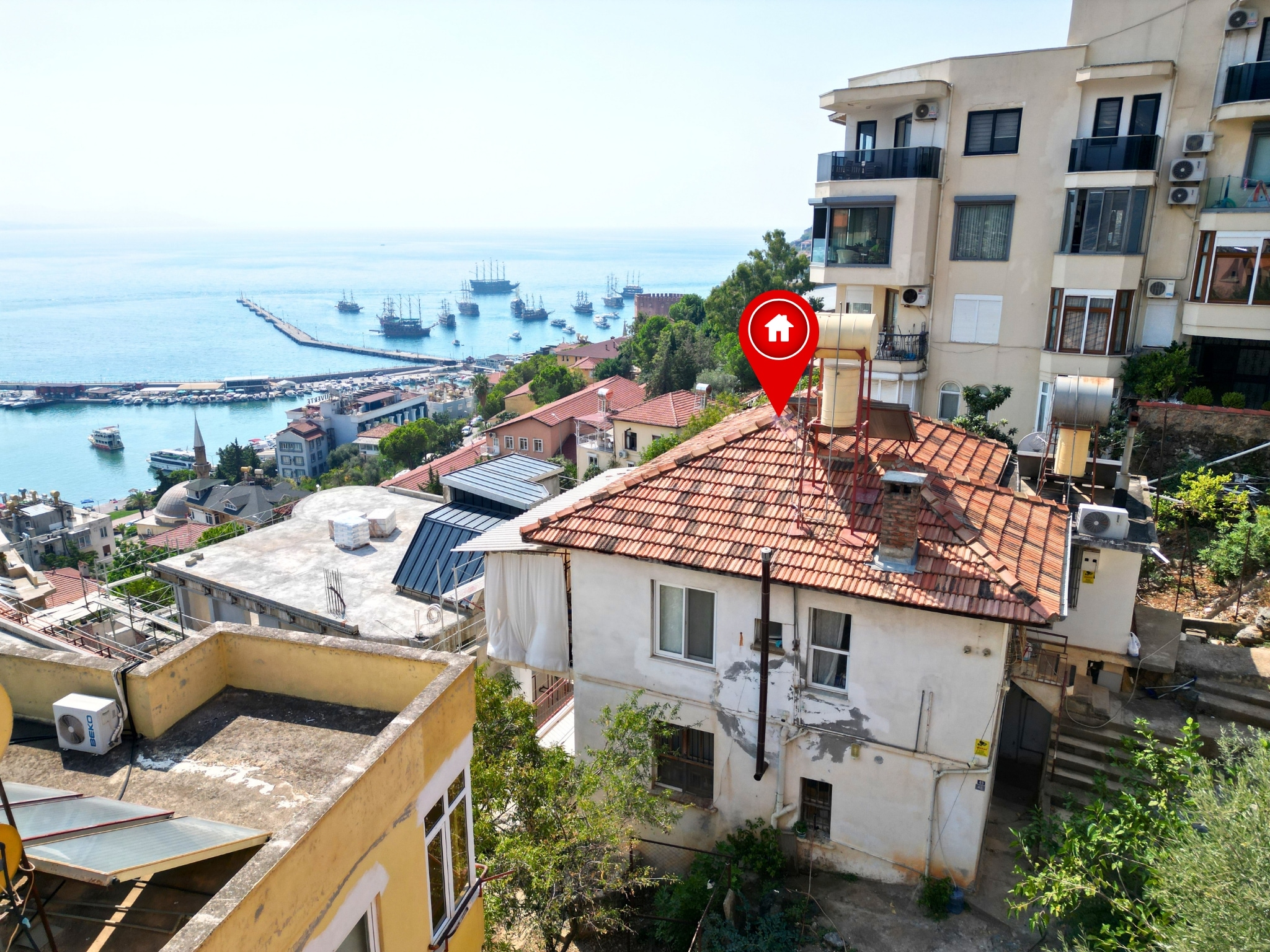 A harbor with piers and boats beside hillside homes frames a terracotta-roofed villa in Turkey