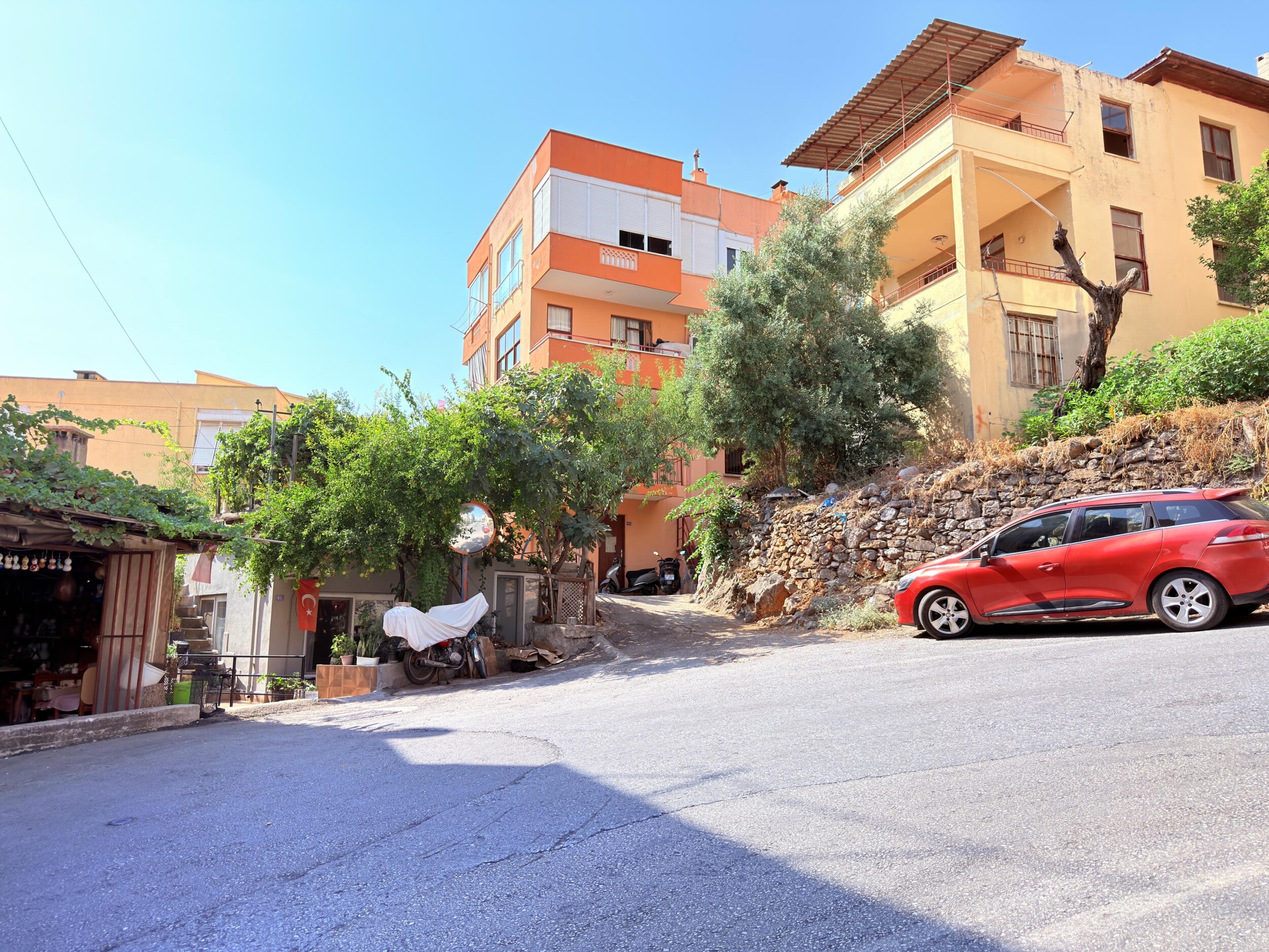 An asphalt street with an orange apartment building and stone retaining wall shows a house for sale in Turkey