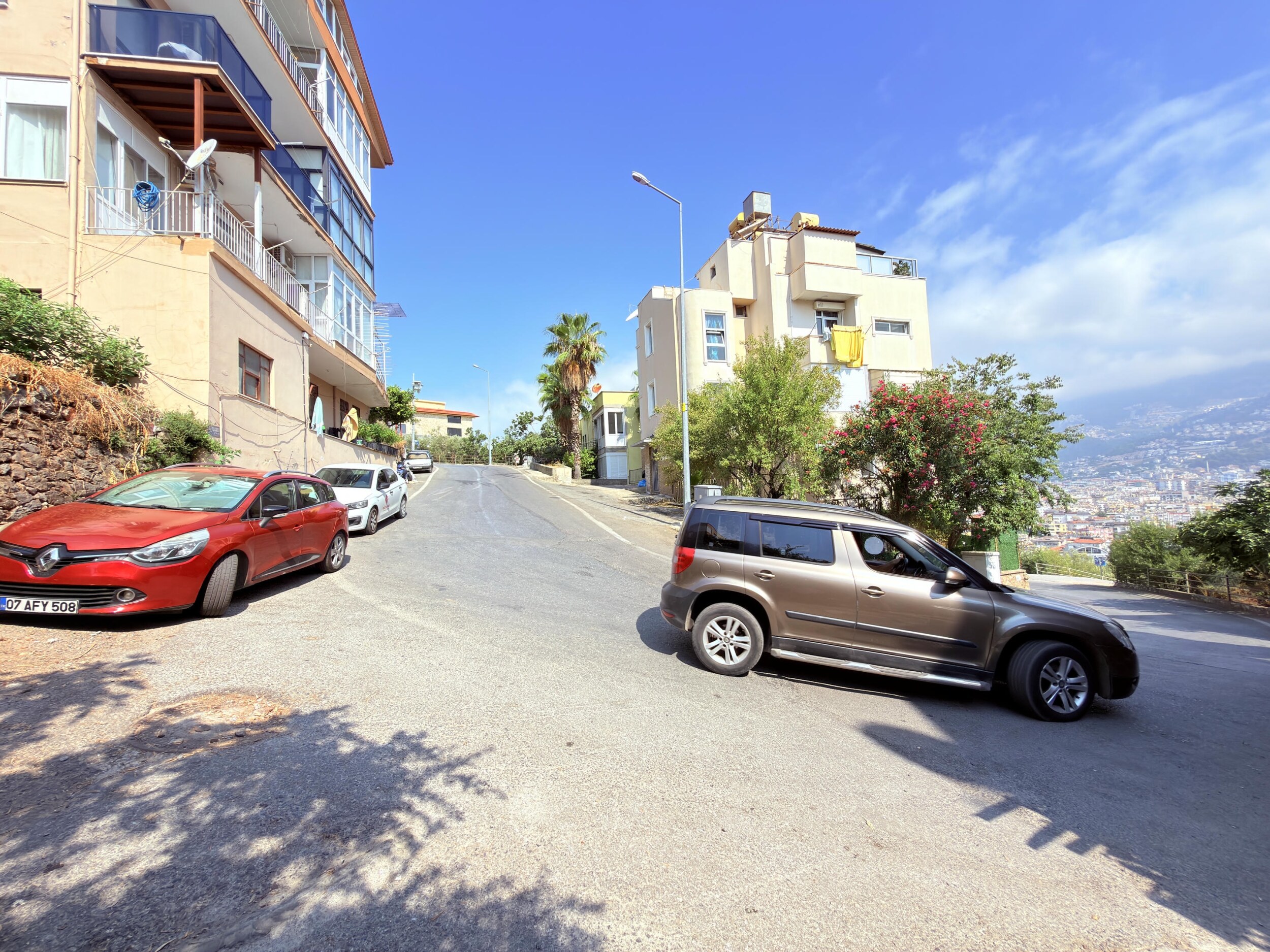 A sloping asphalt street with homes, a palm tree and parked cars passes by a villa