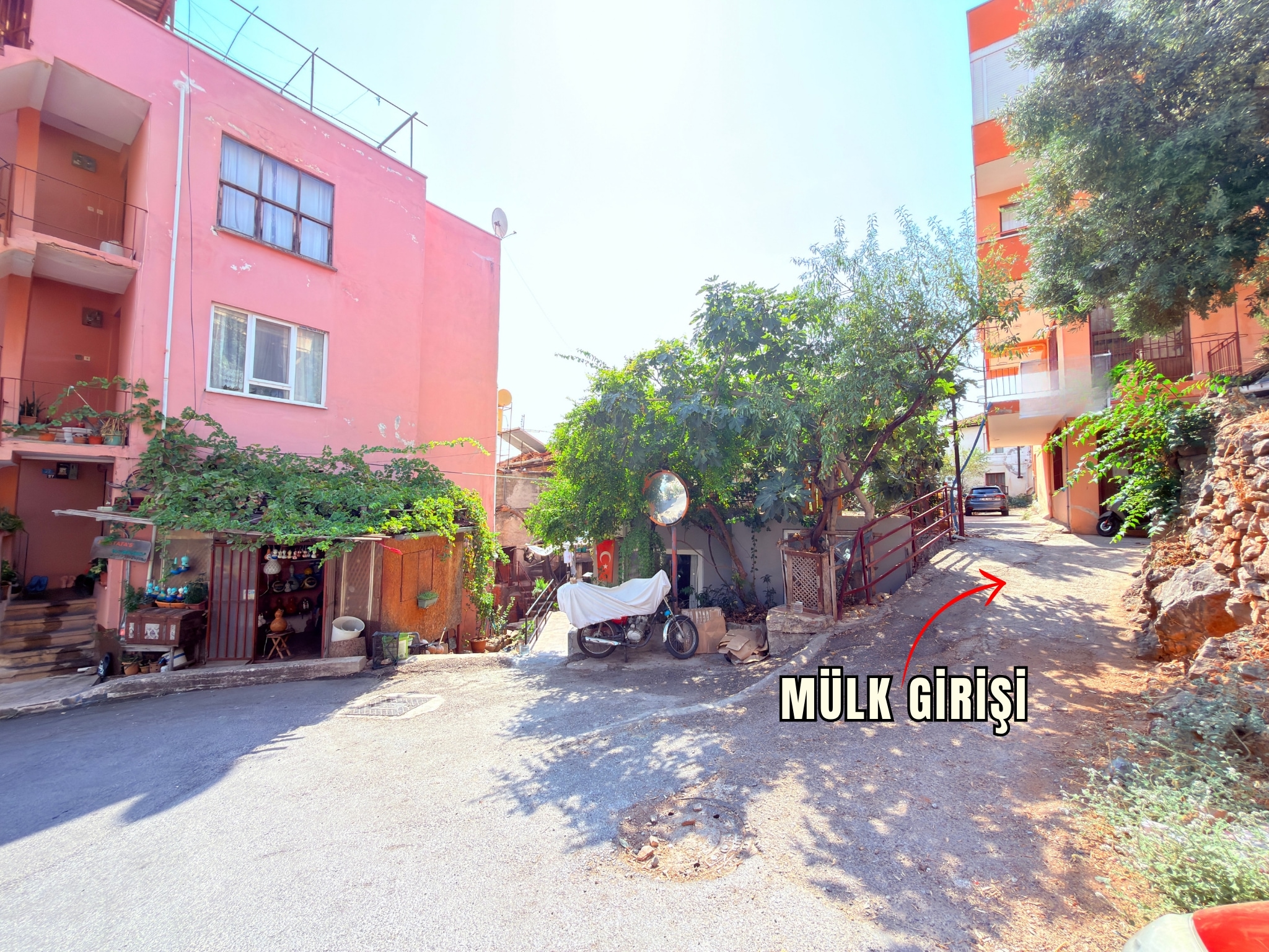A narrow driveway between residential buildings and trees leads to a house for sale