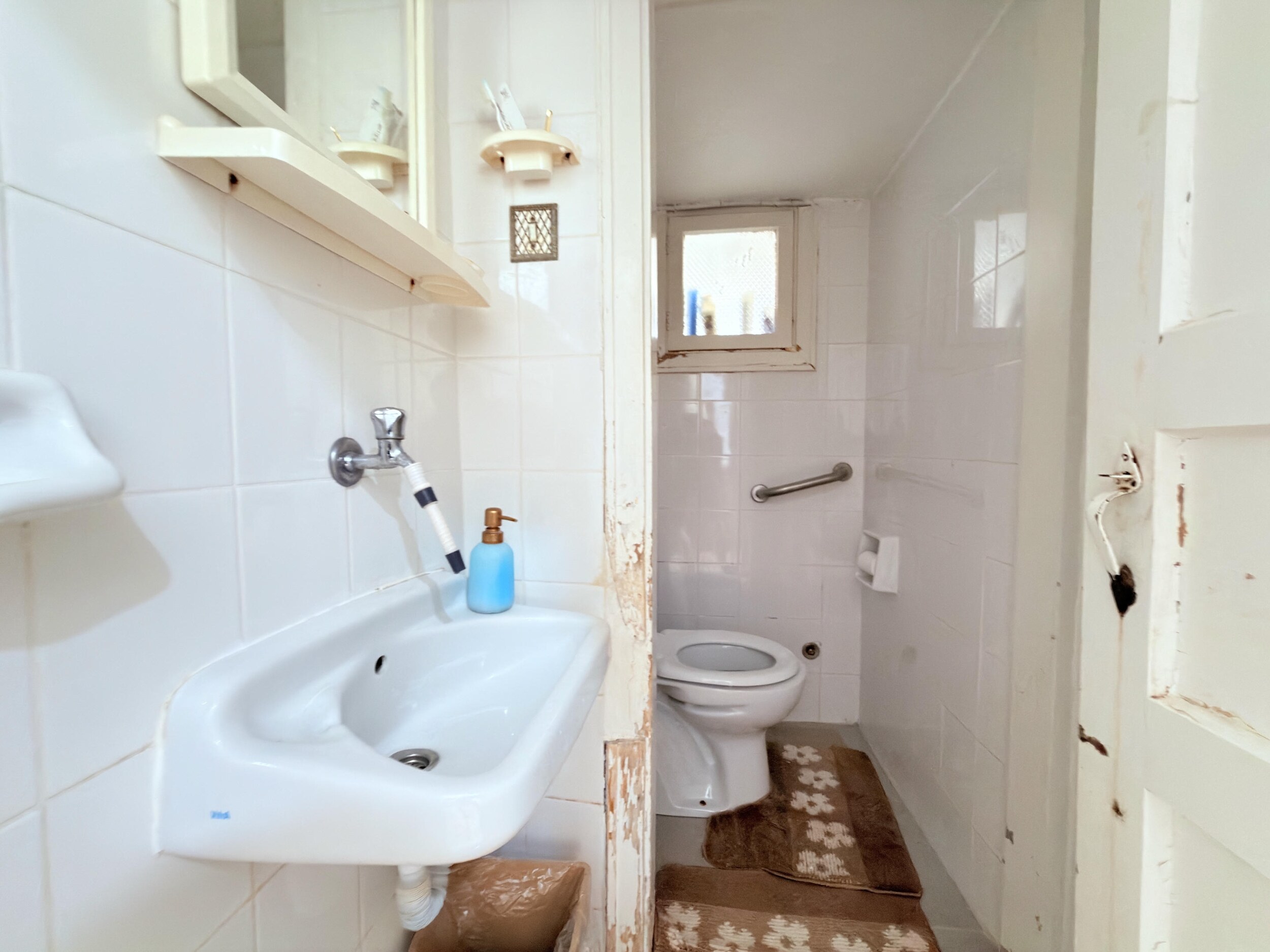 A narrow bathroom with white tiles, wall-mounted sink and small window in Alanya