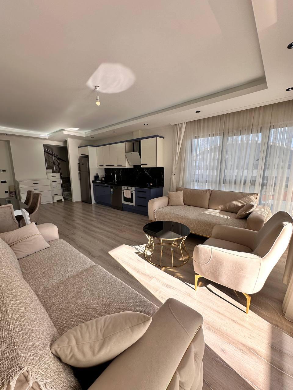 Açık renkli kanepe ve iki koltuk, Mahmutlar’da penthouse salonda panoramik pencereler ve mavi-beyaz mutfak önünde
