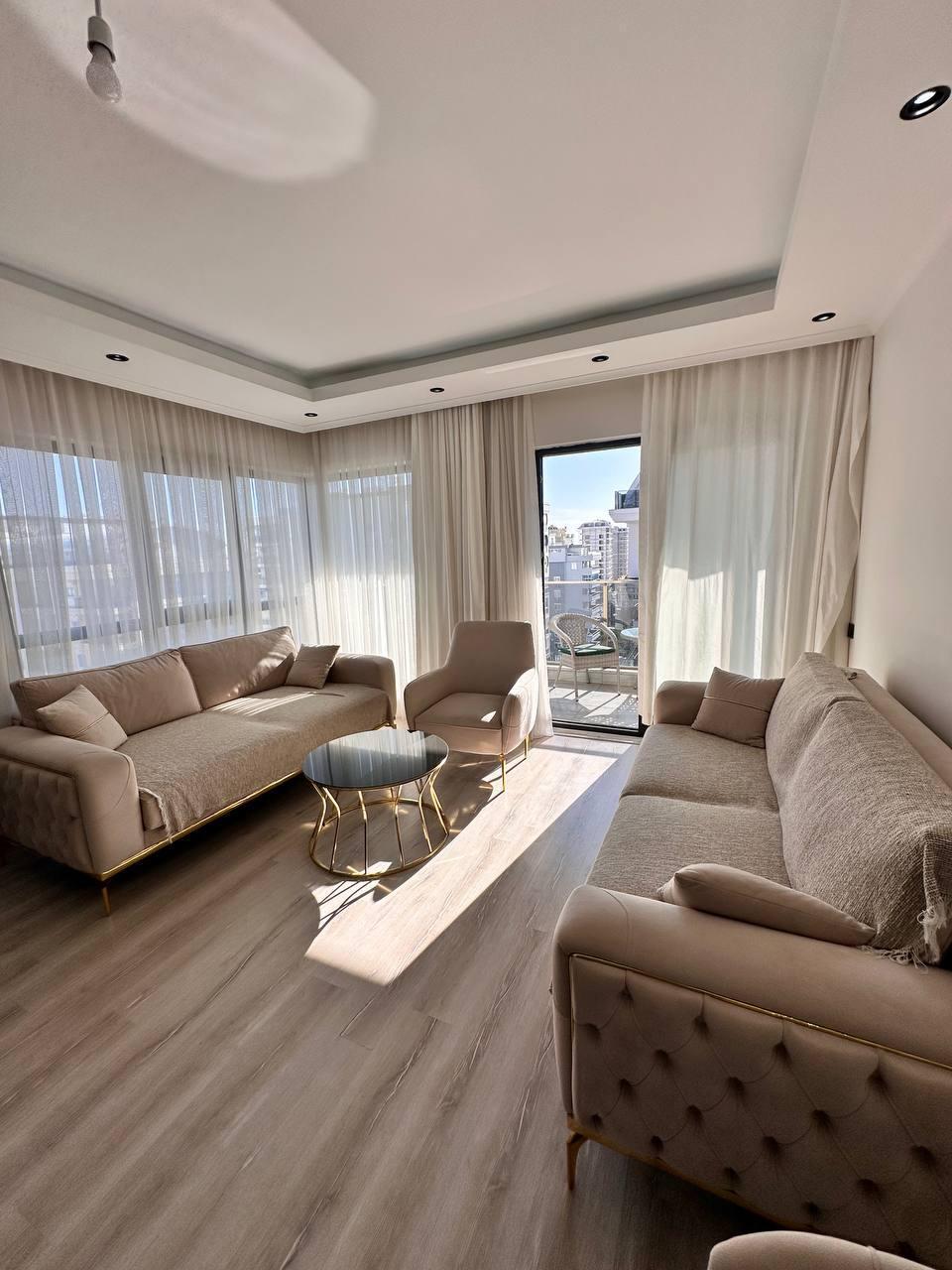 İki bej kanepe ve yuvarlak sehpa, Türkiye’de rezidansta balkon kapısından görünen şehir manzarasına bakıyor