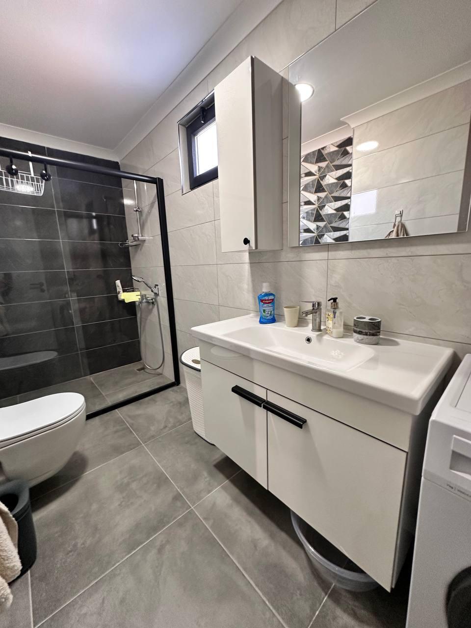 Cam bölmeli duşakabin, penthouse banyosunda lavabo dolabı ve çamaşır makinesiyle birlikte görülüyor