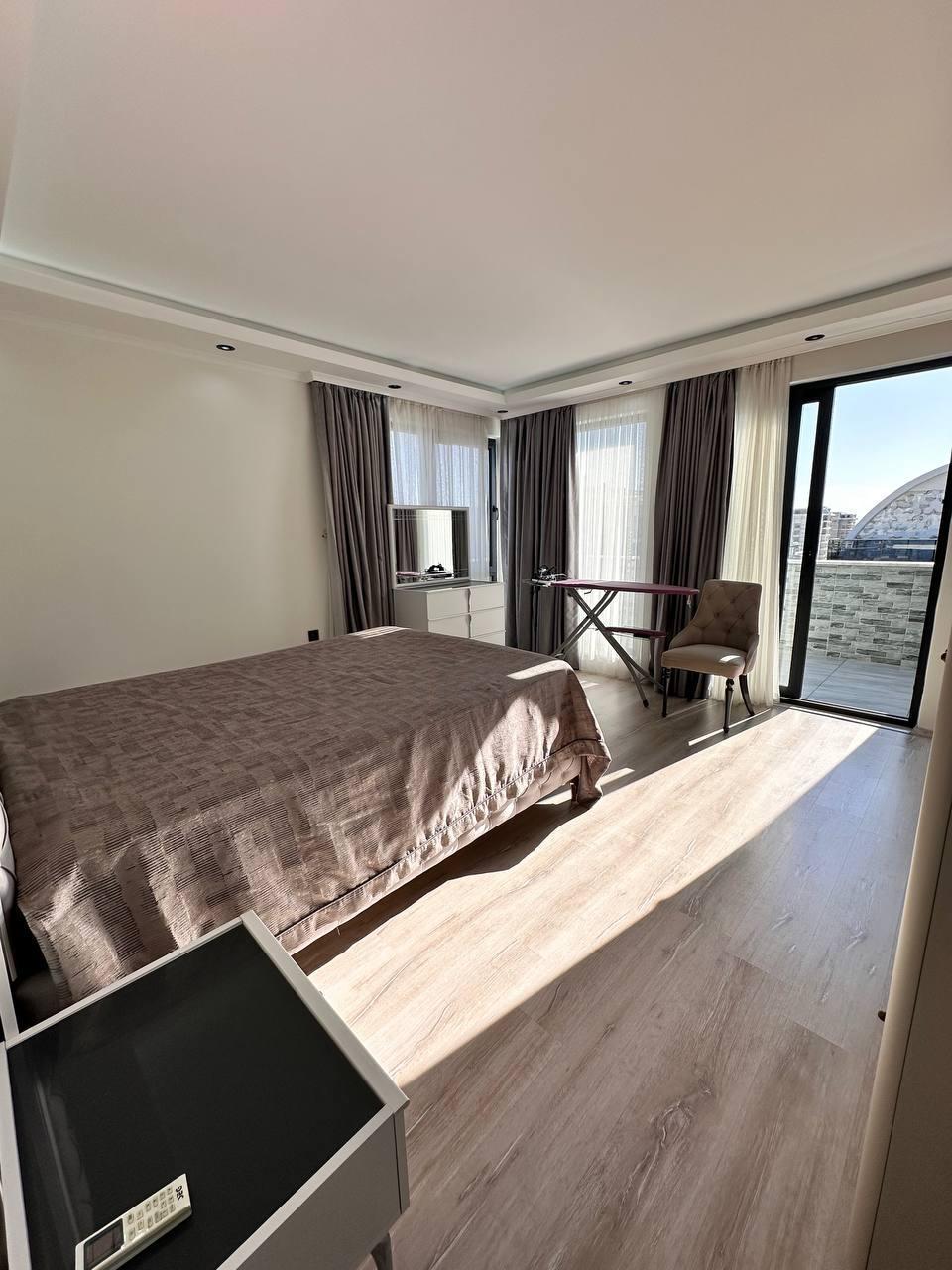 Kahverengi örtülü yatak, penthouse yatak odasında terasa açılan cam kapı ve pencere yanındaki çalışma masasıyla