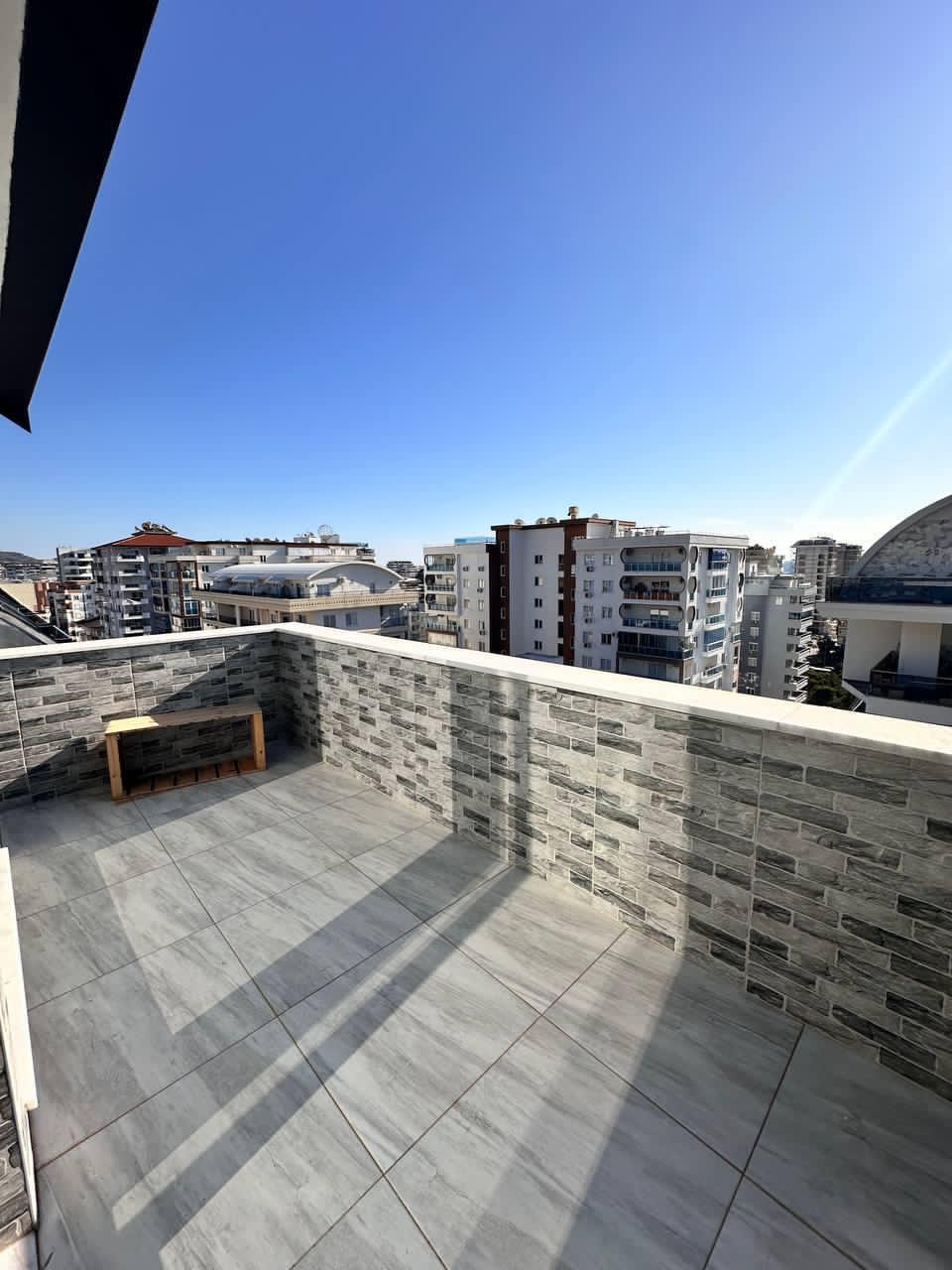 Geniş gri teras, Mahmutlar’da Türkiye’de penthouse yanında şehir manzarası ve duvar kenarında ahşap bankla