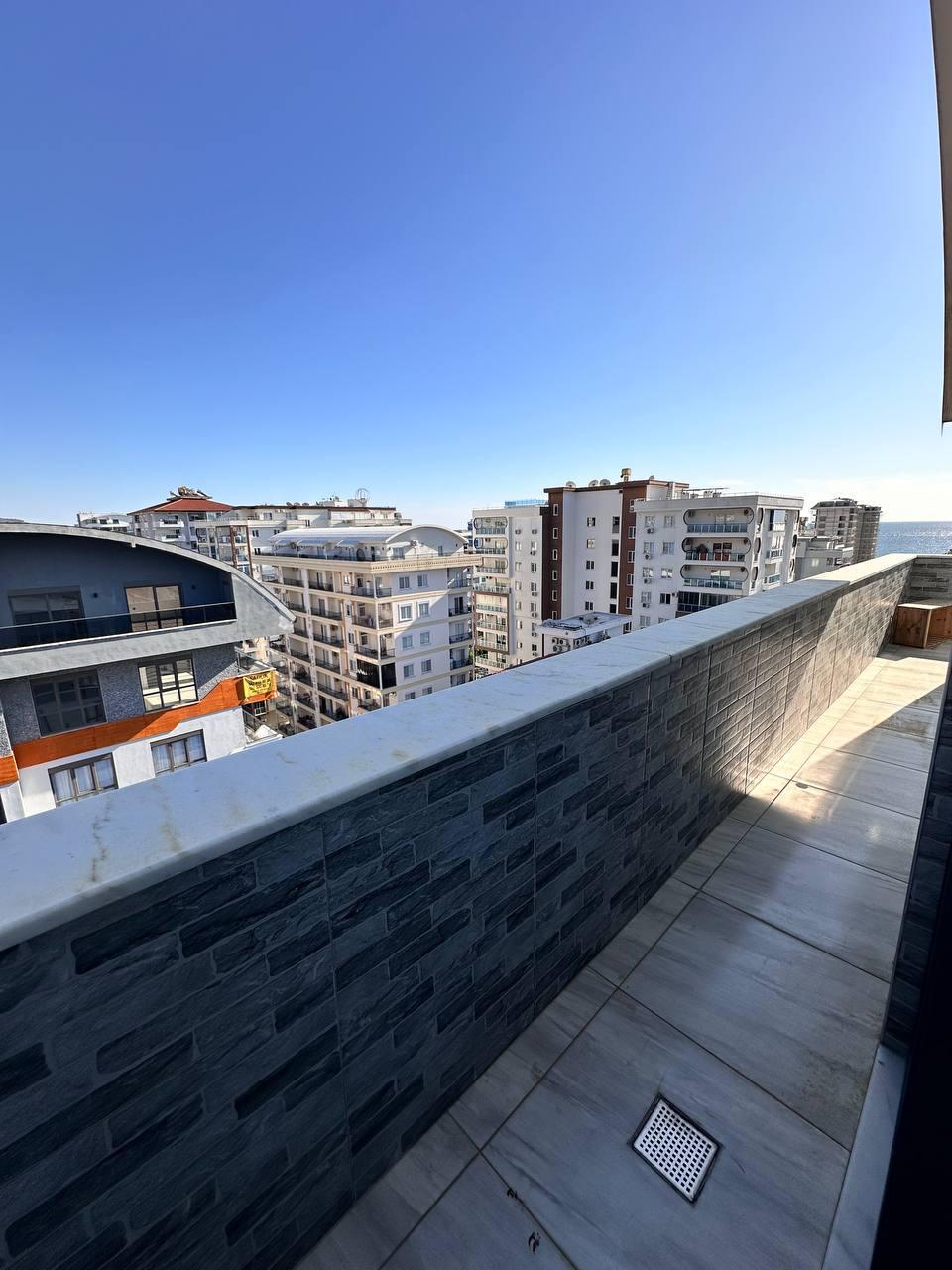 Uzun taş korkuluklu teras, Mahmutlar’da satışta olan penthouse için deniz manzarası ve şehir binalarıyla açılıyor