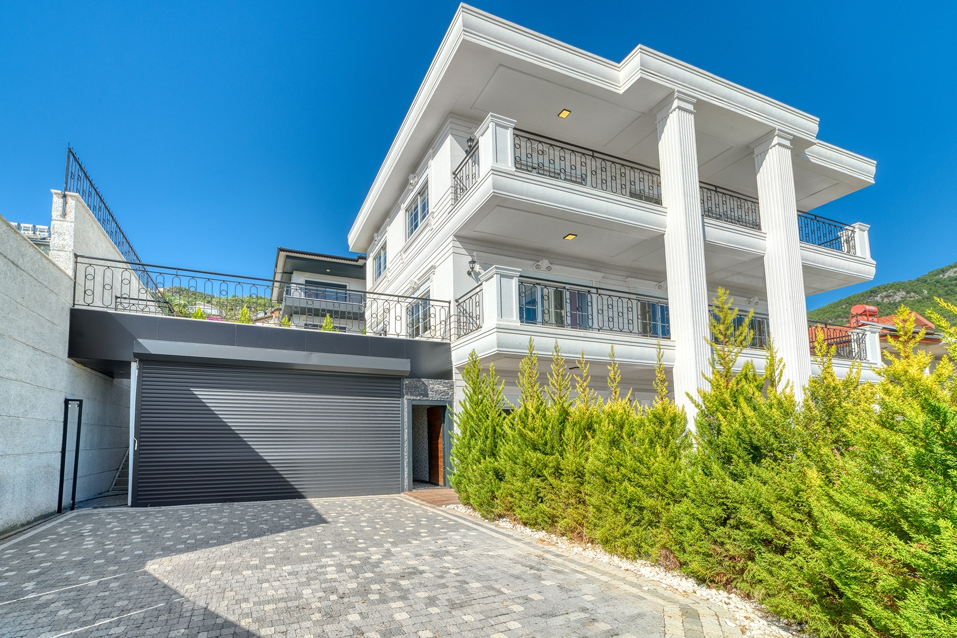 White villa facade with three balcony levels and a closed garage against a mountain slope in Turkey