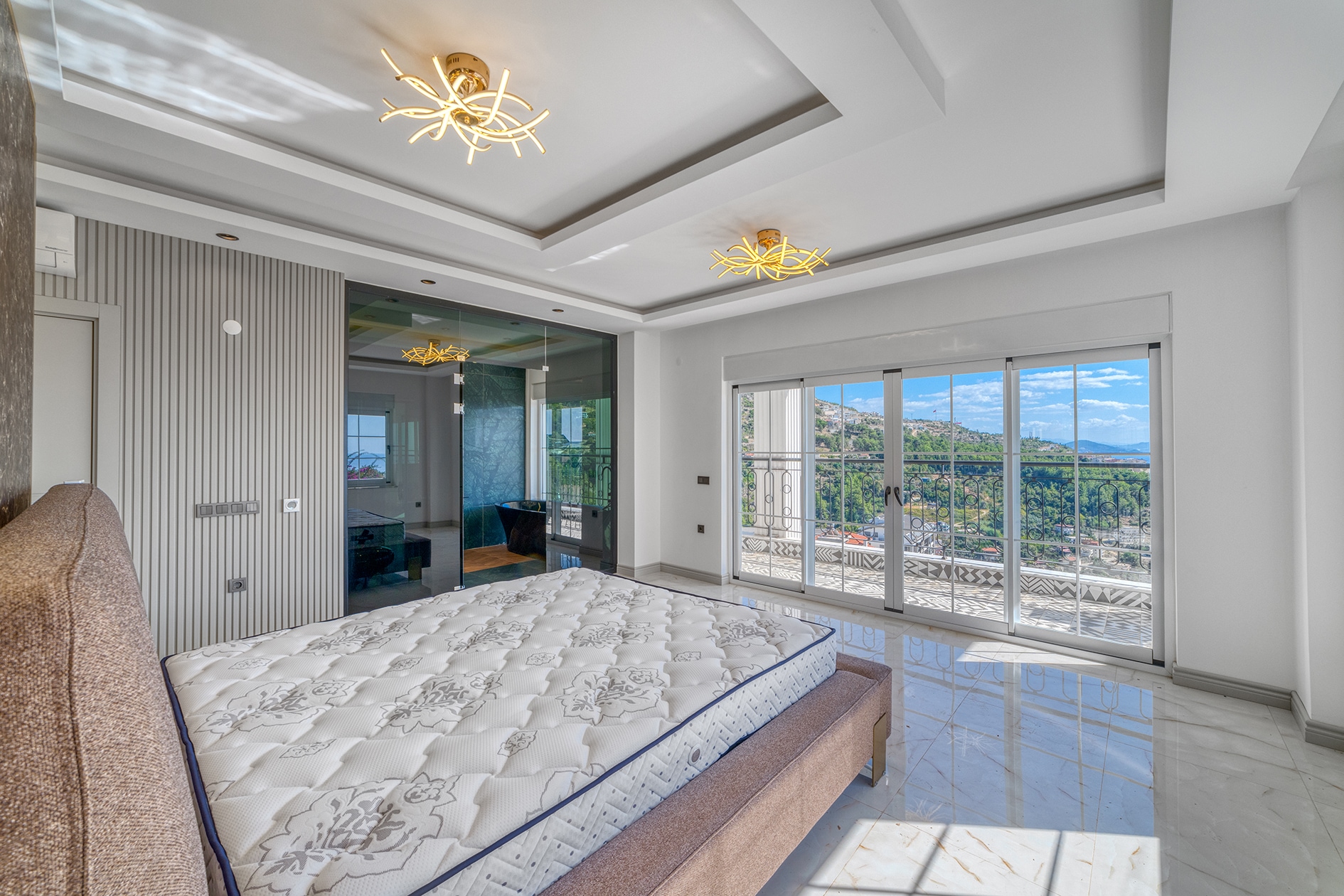 Beige bed by balcony doors with views of green hills and city buildings in a villa in Alanya