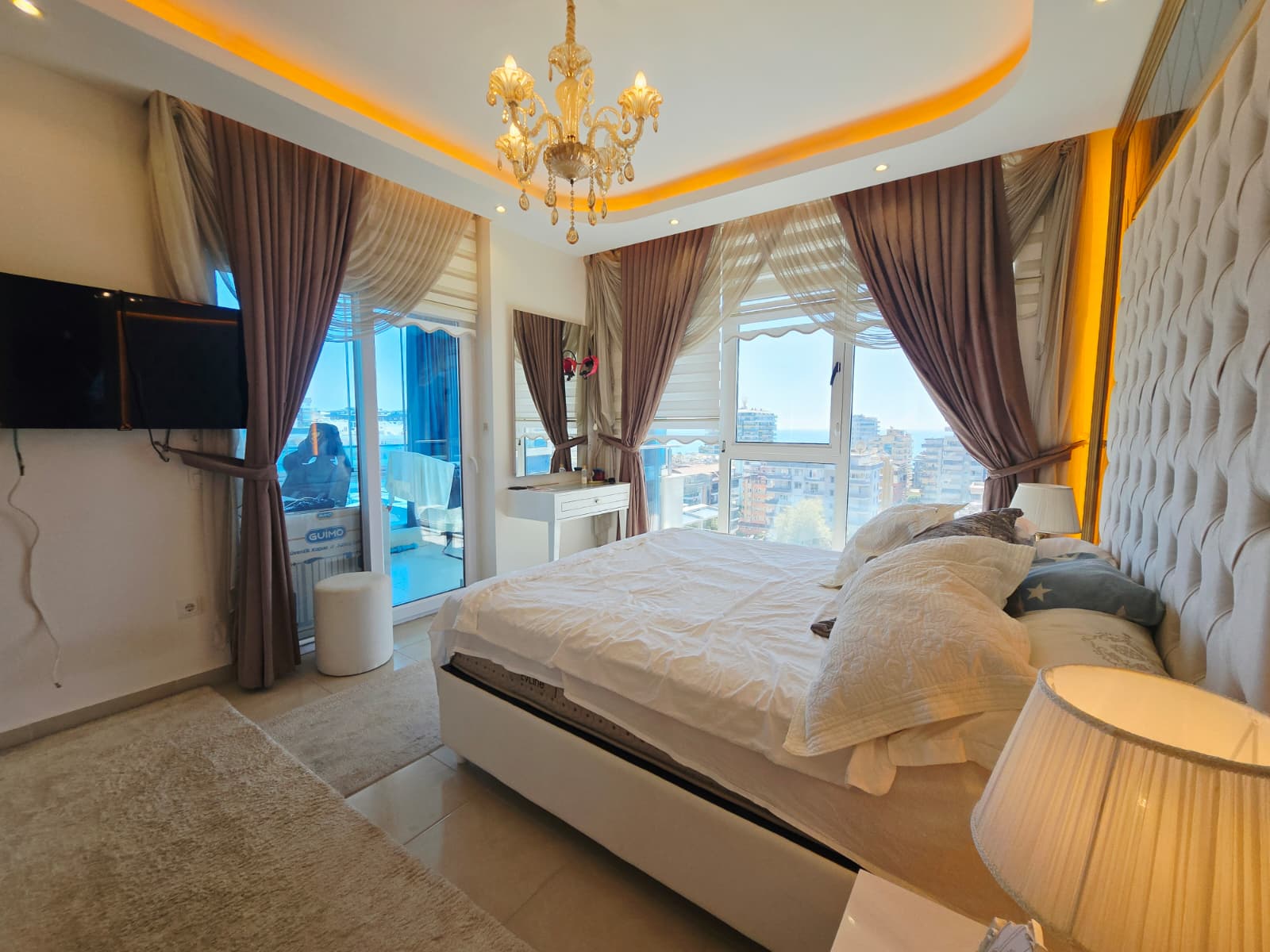 Beyaz örtülü çift kişilik yatak, şehir manzaralı panoramik pencereler ve kristal avize çatı katı dairede