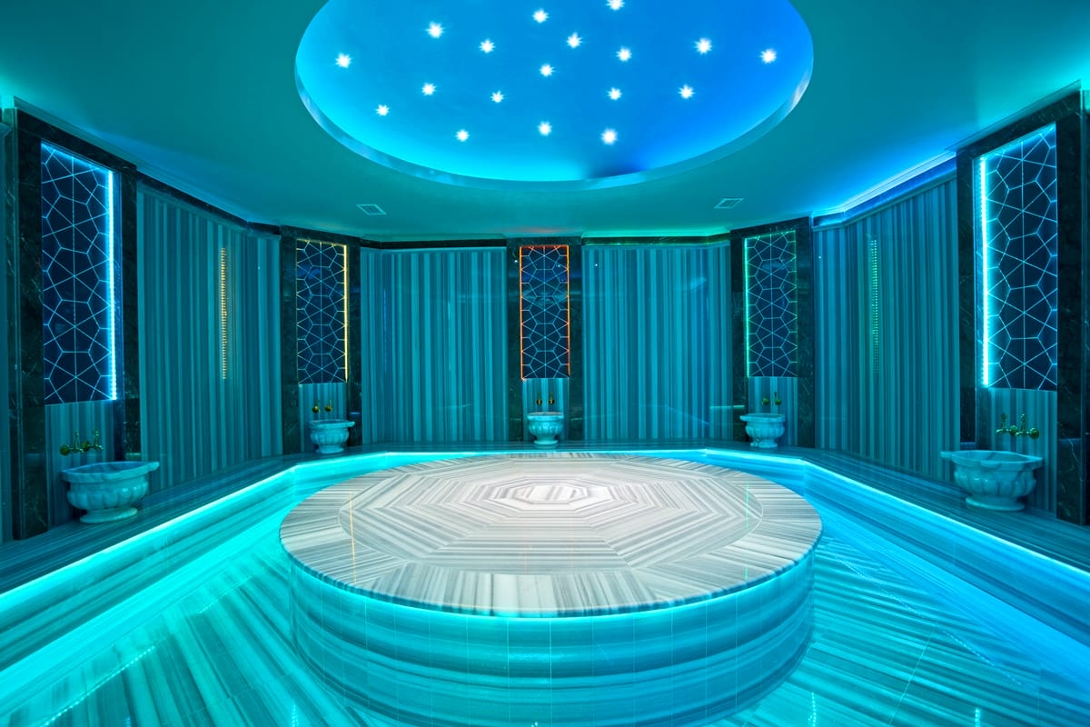 Hammam with a round marble platform, mosaic surfaces and blue accent lighting in a complex with houses