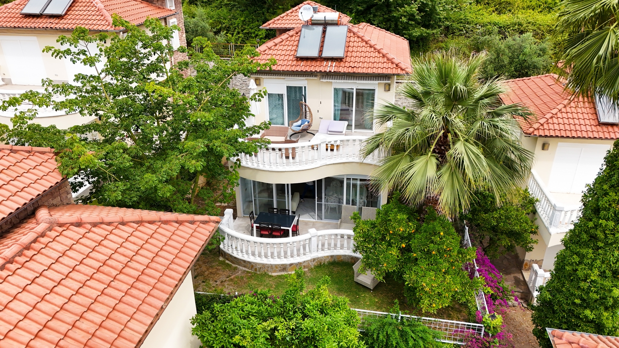Red-roofed villa with solar panels and curved terraces surrounded by palms and fruit trees in Alanya, Turkey