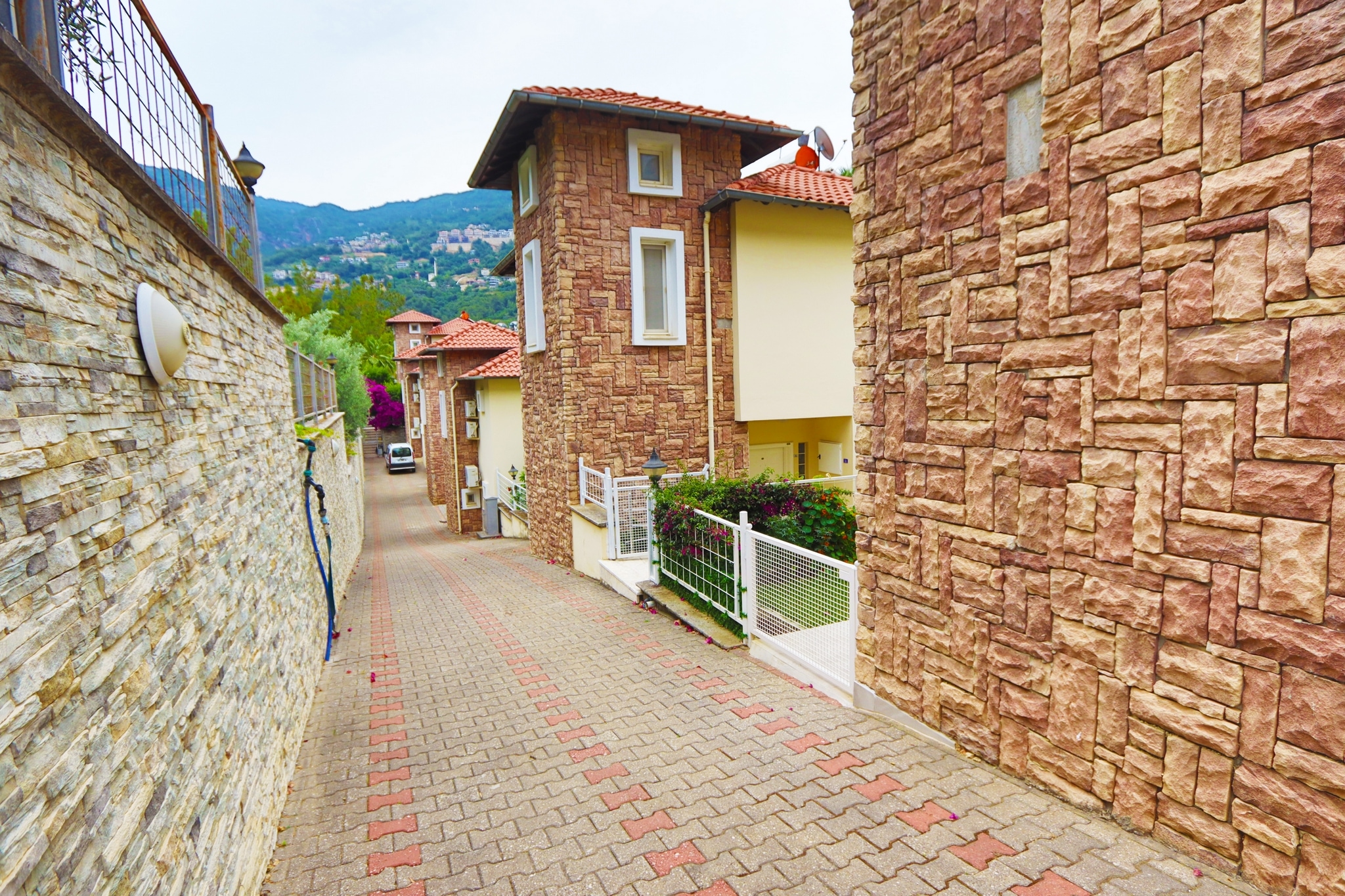 Paved driveway with stone retaining walls, red-roofed homes and mountain slopes in Turkey where it is for sale