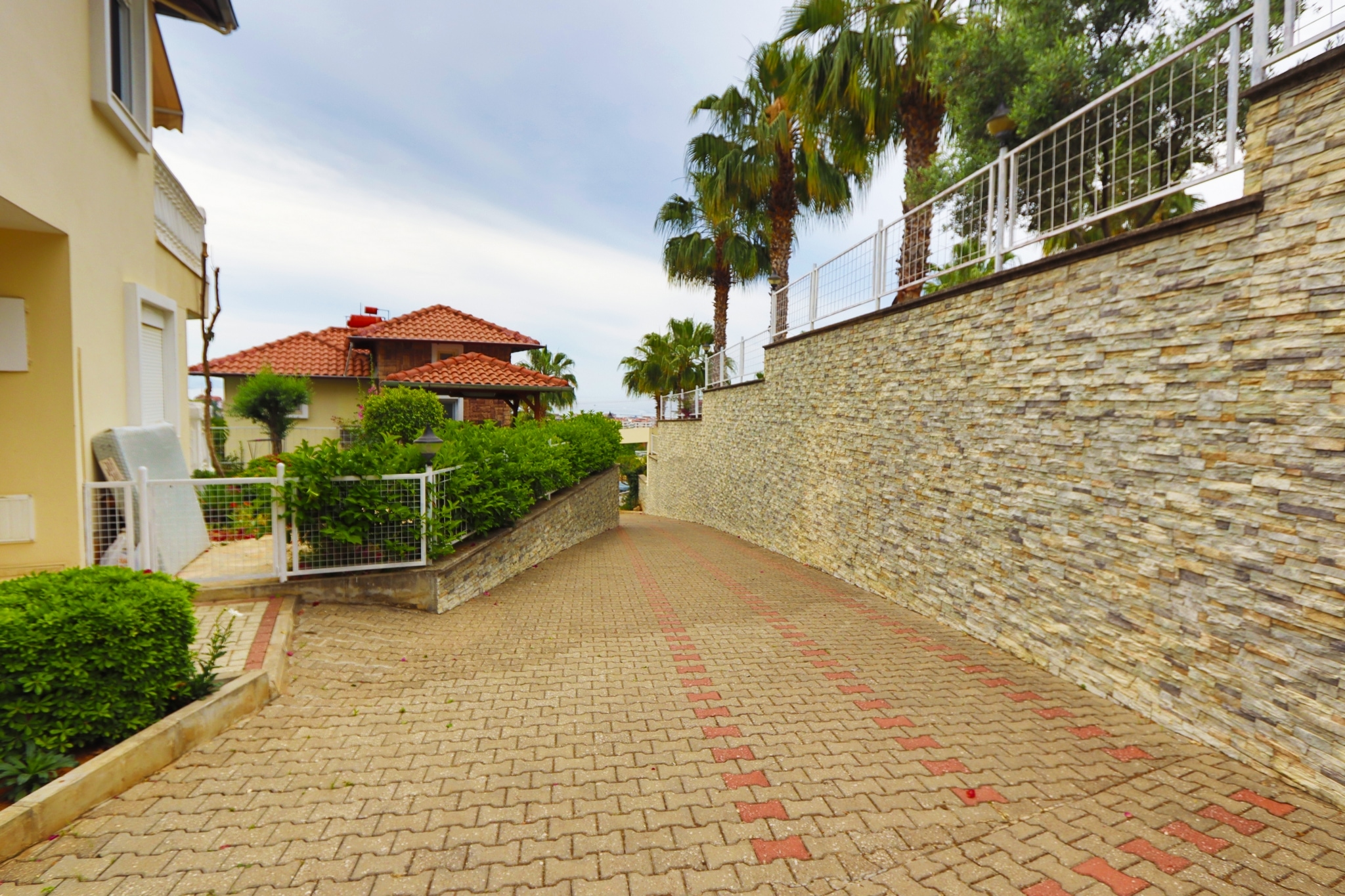 Wide paved path with palms, white gates and neighboring red-tiled roofs beside a villa