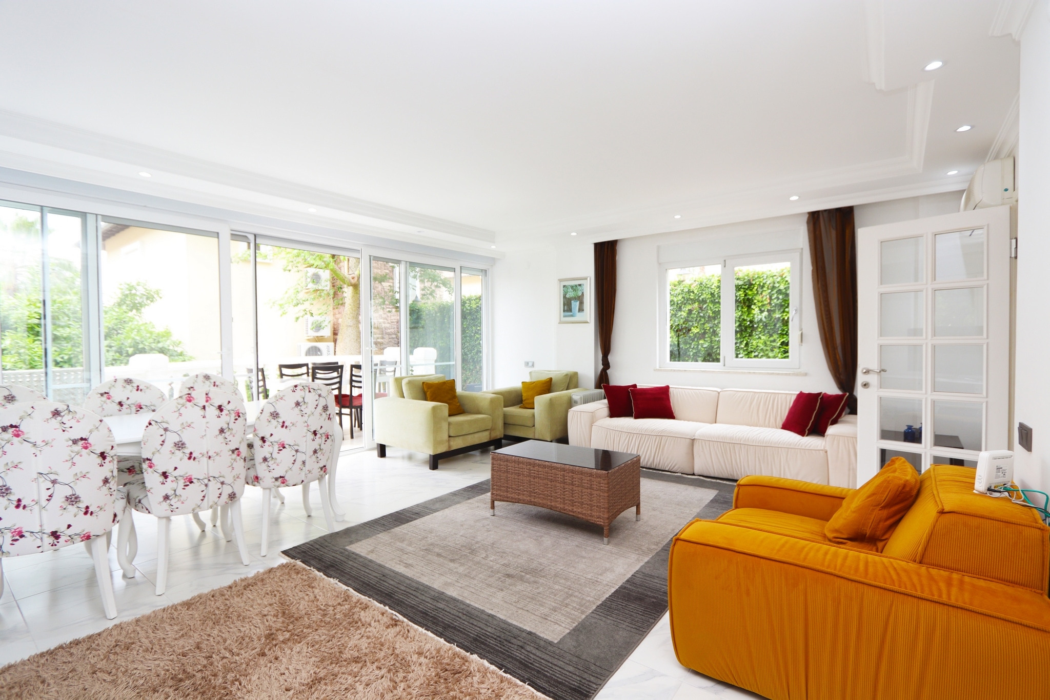 Kitchen-living room with white and orange sofas, panoramic glazing and greenery outside in a villa for sale