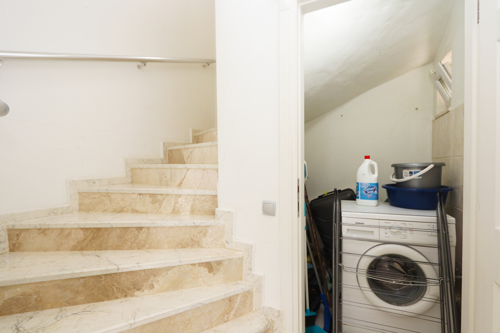 Laundry room beneath a staircase with marble steps, washing machine and folding drying rack