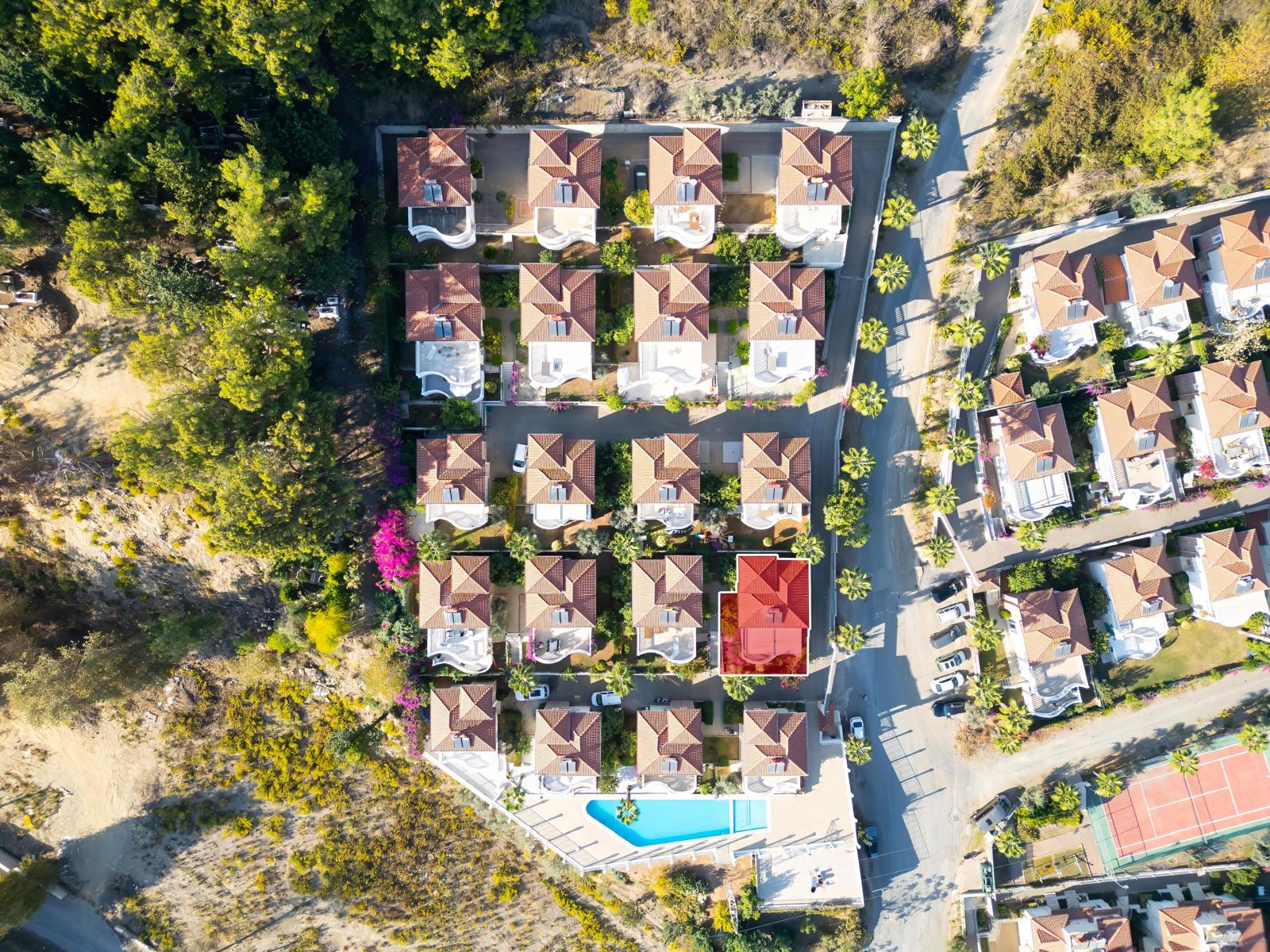 Aerial view of a villa complex with red roofs, a shared pool, tennis court and wooded surroundings