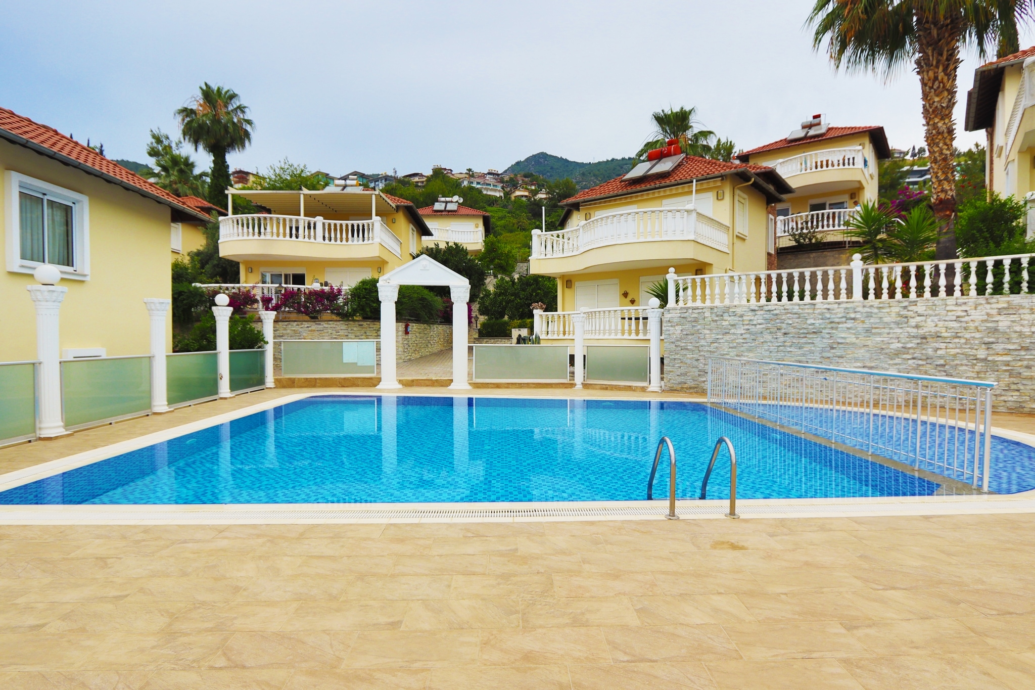 Rectangular outdoor pool with a metal ladder, palms and green hills beside a villa in Alanya