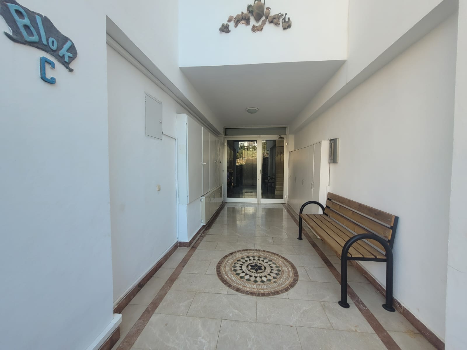 Narrow shared corridor with a wooden bench, mosaic floor inlay and glass entrance doors