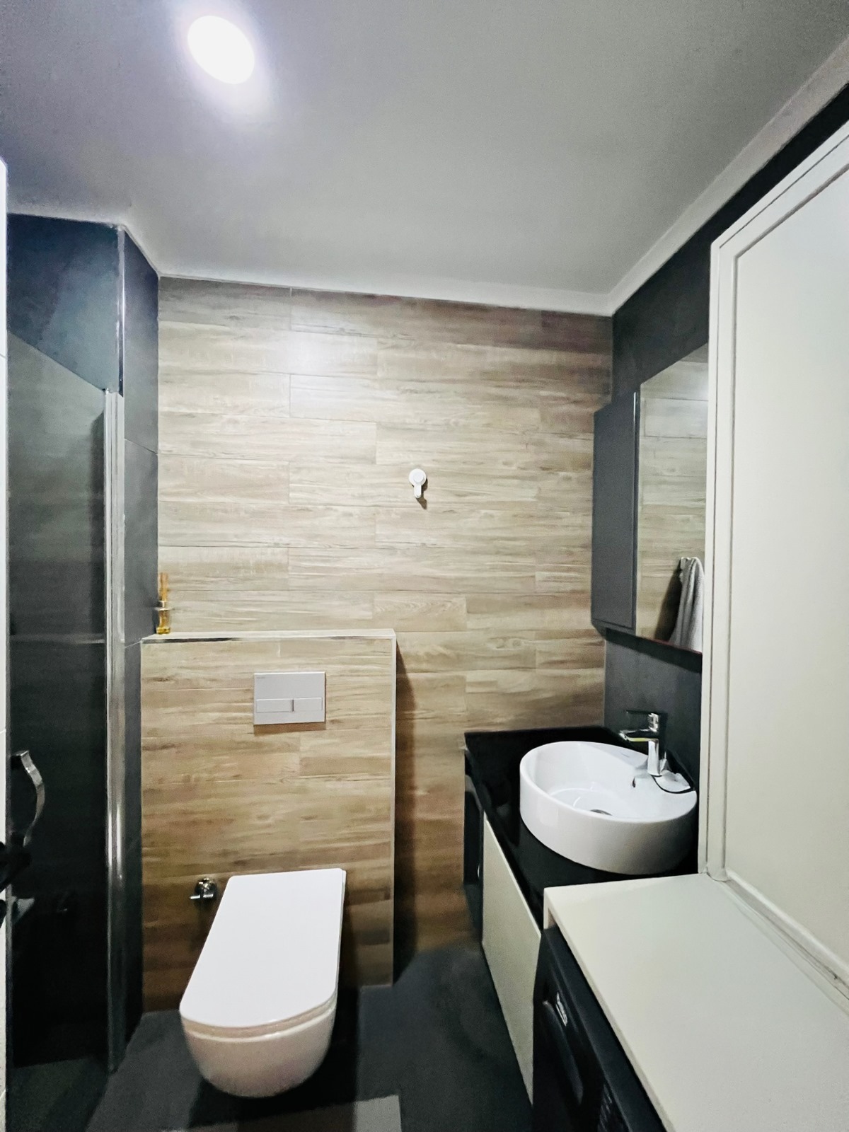 Cam duş kabinli banyo, büyük ayna ve ahşap görünümlü duvar kaplamasıyla düzenlenmiş