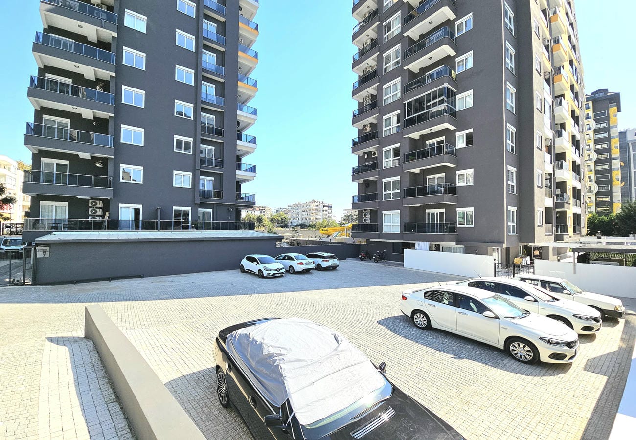 Open parking with cars beside dark gray residential towers at a residence in Mahmutlar