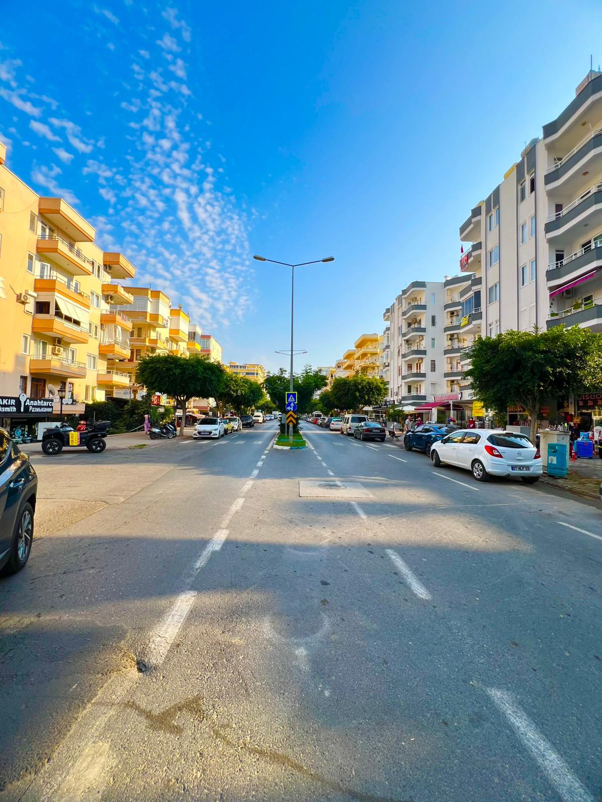 Mahmutlar’da mağazalı yüksek binalar ve park etmiş araçlarla geniş şehir caddesi, Türkiye
