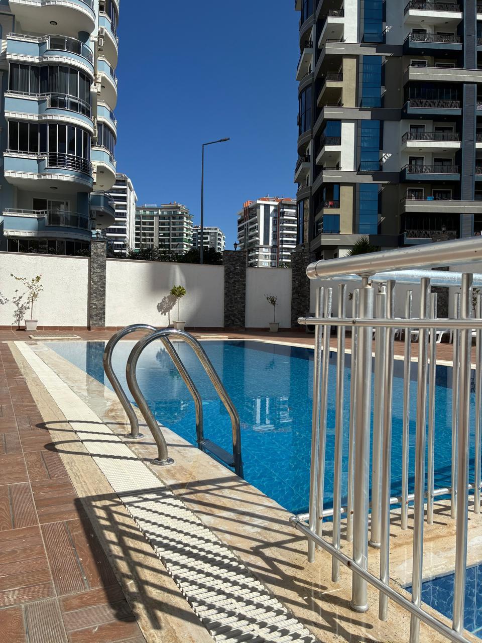 Outdoor pool with a safety barrier and white perimeter fence among multi-storey homes in Alanya, Turkey