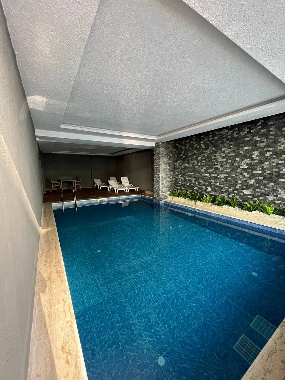 Long indoor pool with blue mosaic lining and a planted bed by a dark stone wall in a residence in Mahmutlar