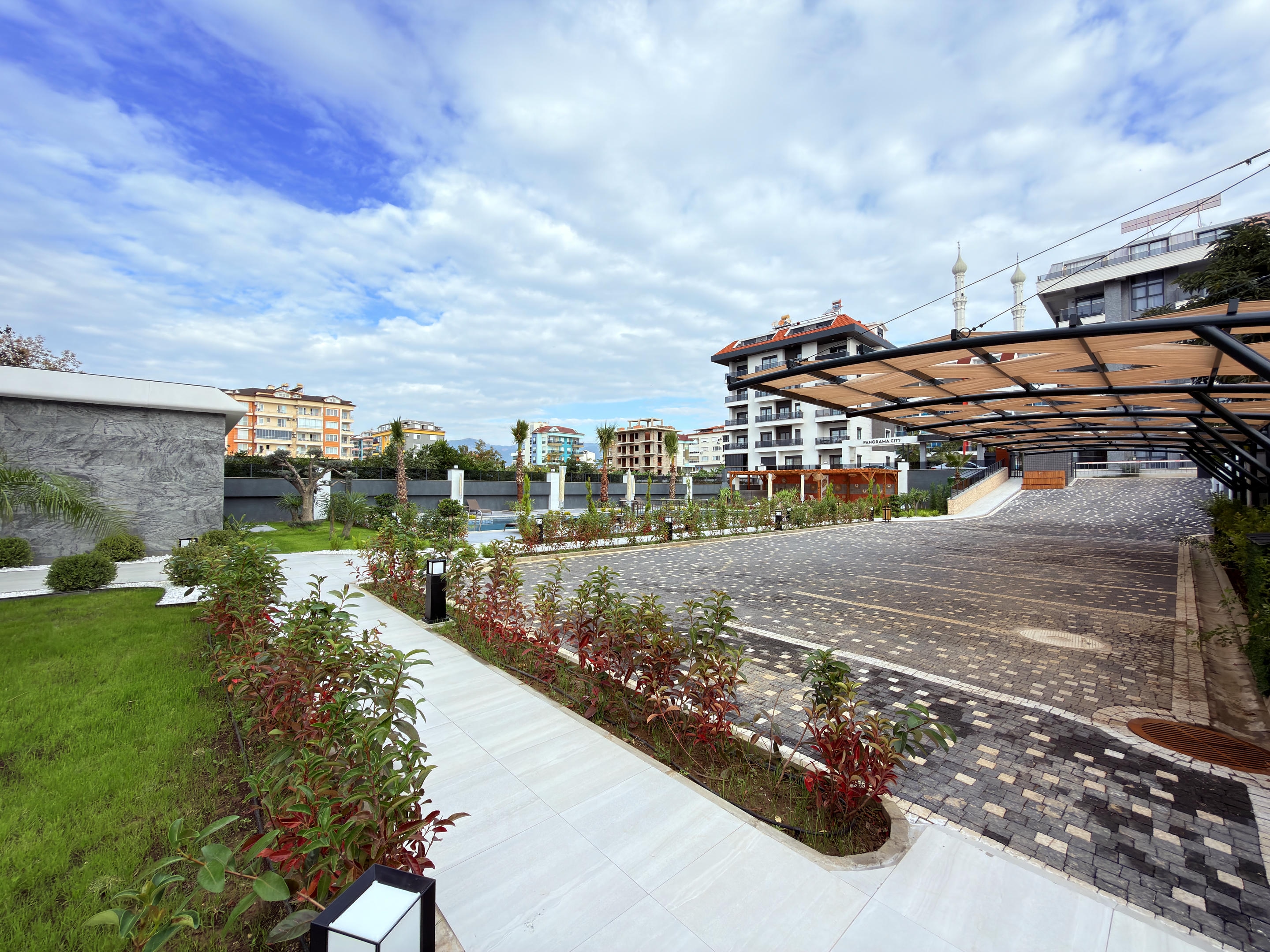 Paved parking area beneath a long carport with planted beds and a pool in the background