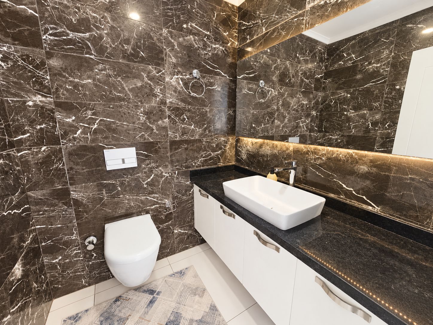 Washroom with dark marble tiles, vessel sink and illuminated wall-wide mirror in a flat in Kargicak