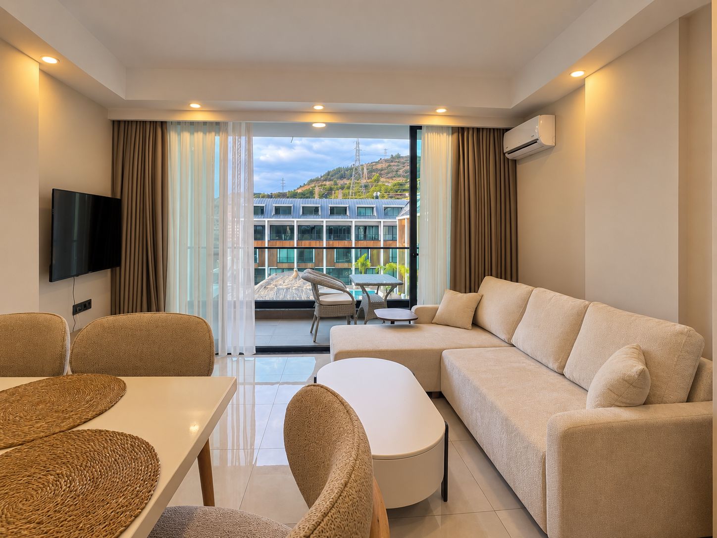Mutfaklı salonda denize yakın satılık konuta ait köşe kanepe, duvar televizyonu ve balkon