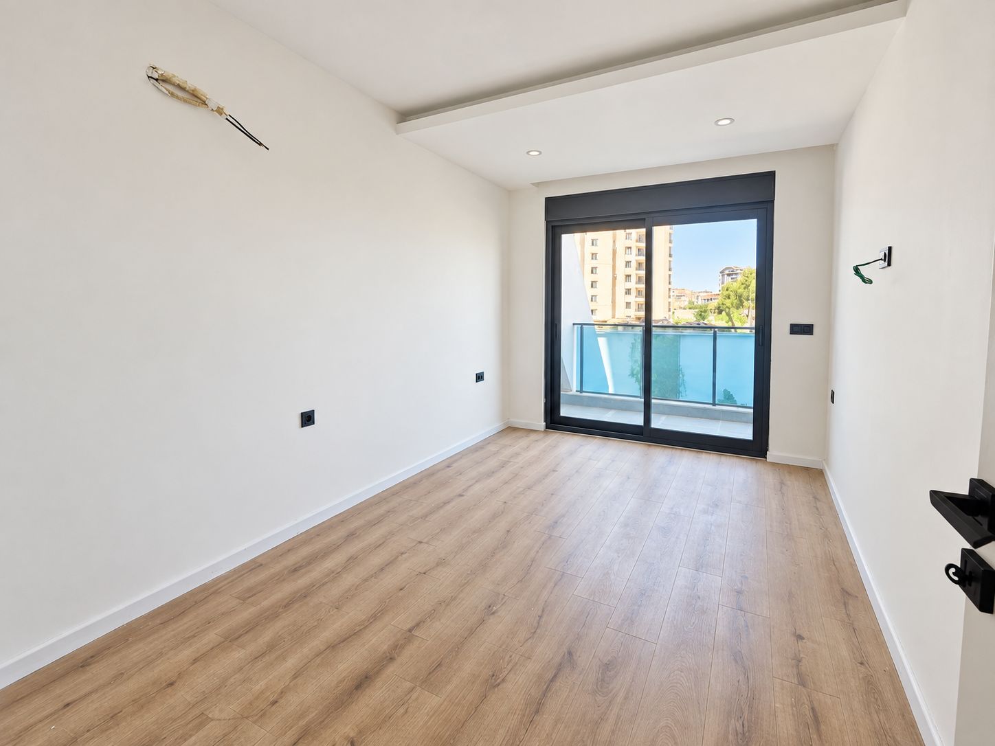 Dairenin boş yatak odasında koyu çerçeveli sürme kapı, mavi cam balkon korkuluğu ve tavan spotları