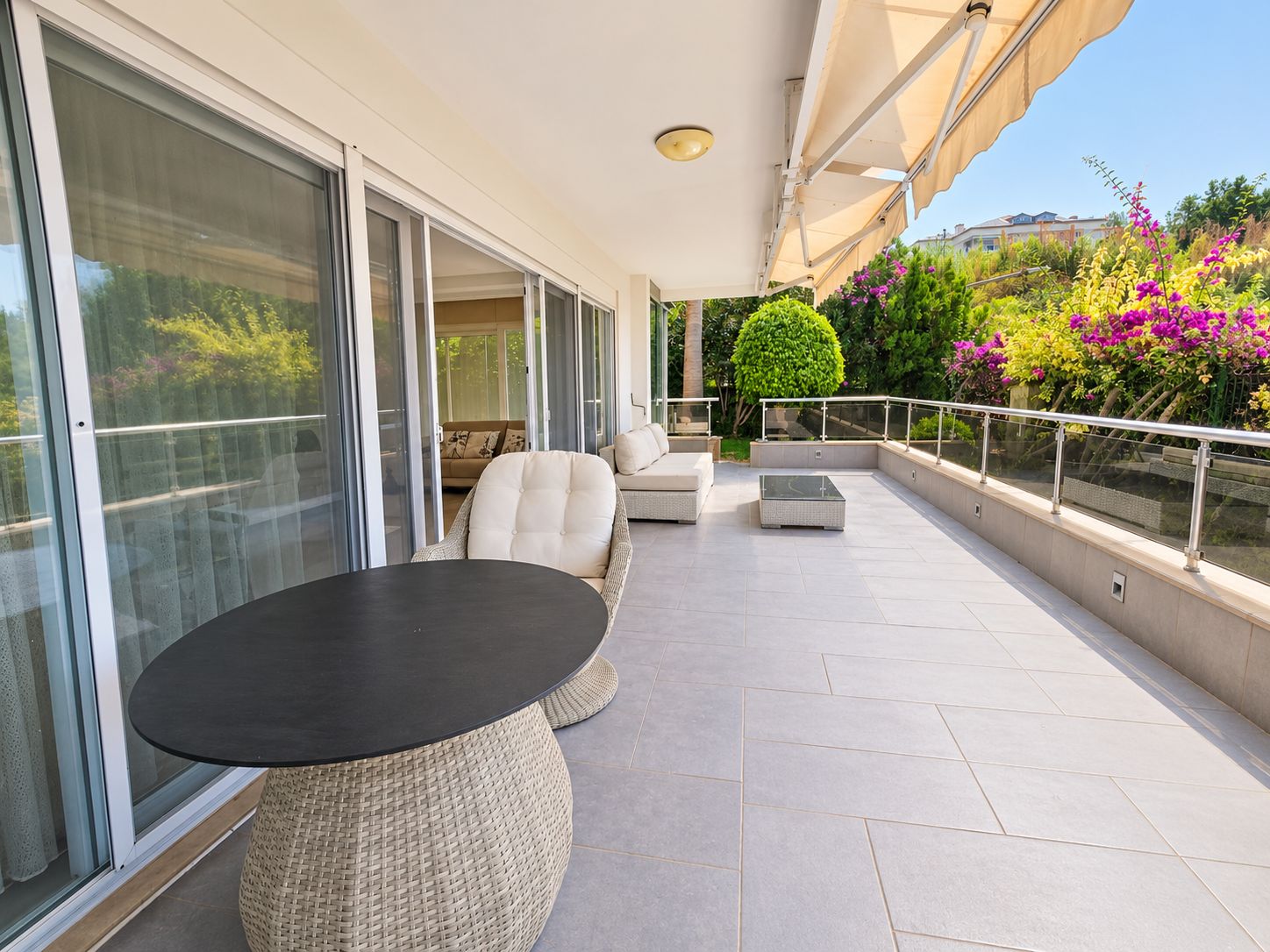 Open terrace with wicker chairs, a round black table and flowering shrubs at a flat for sale