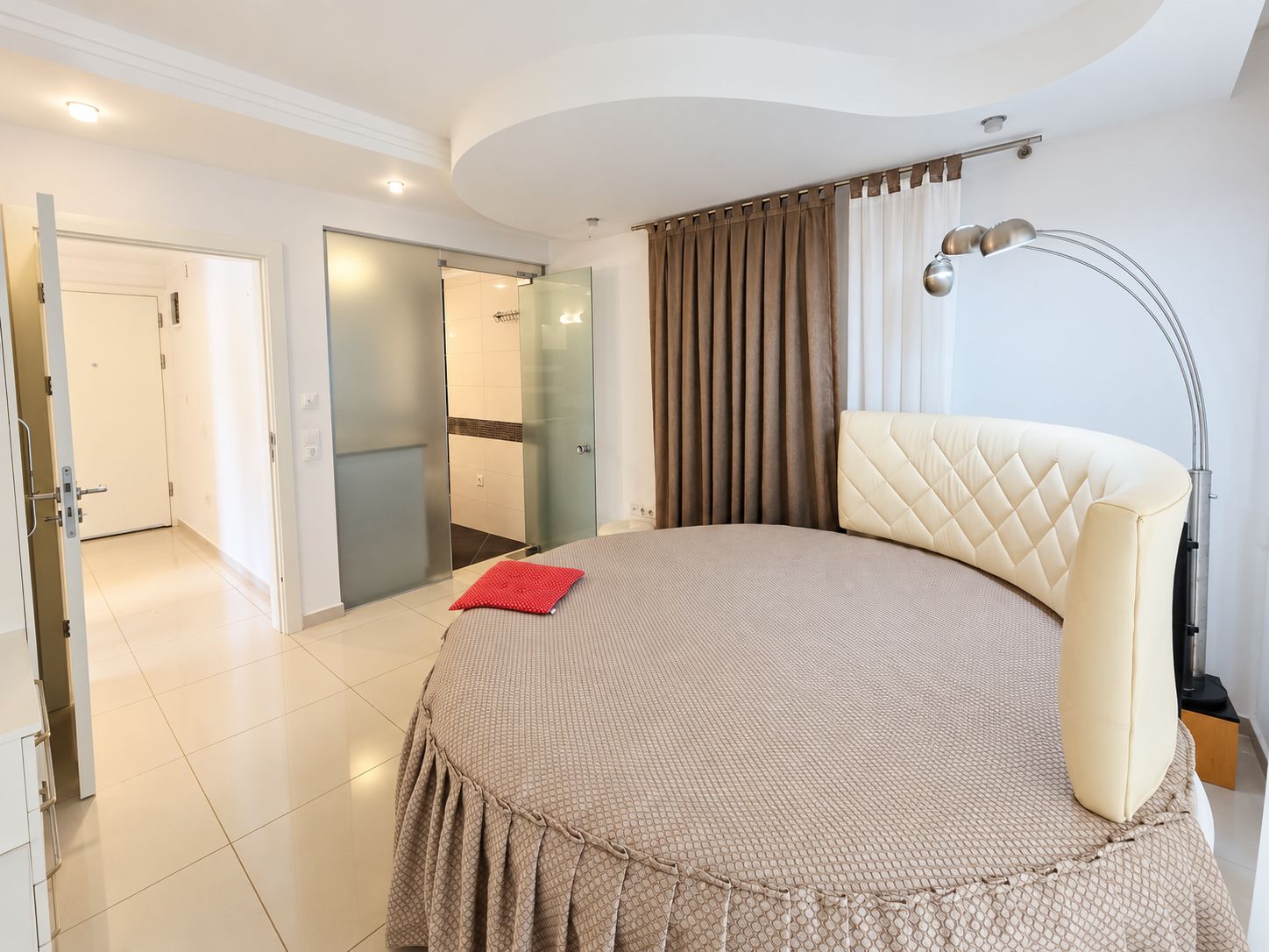 Bedroom with a round double bed, tall beige curtains and bathroom sliding door in Konakli