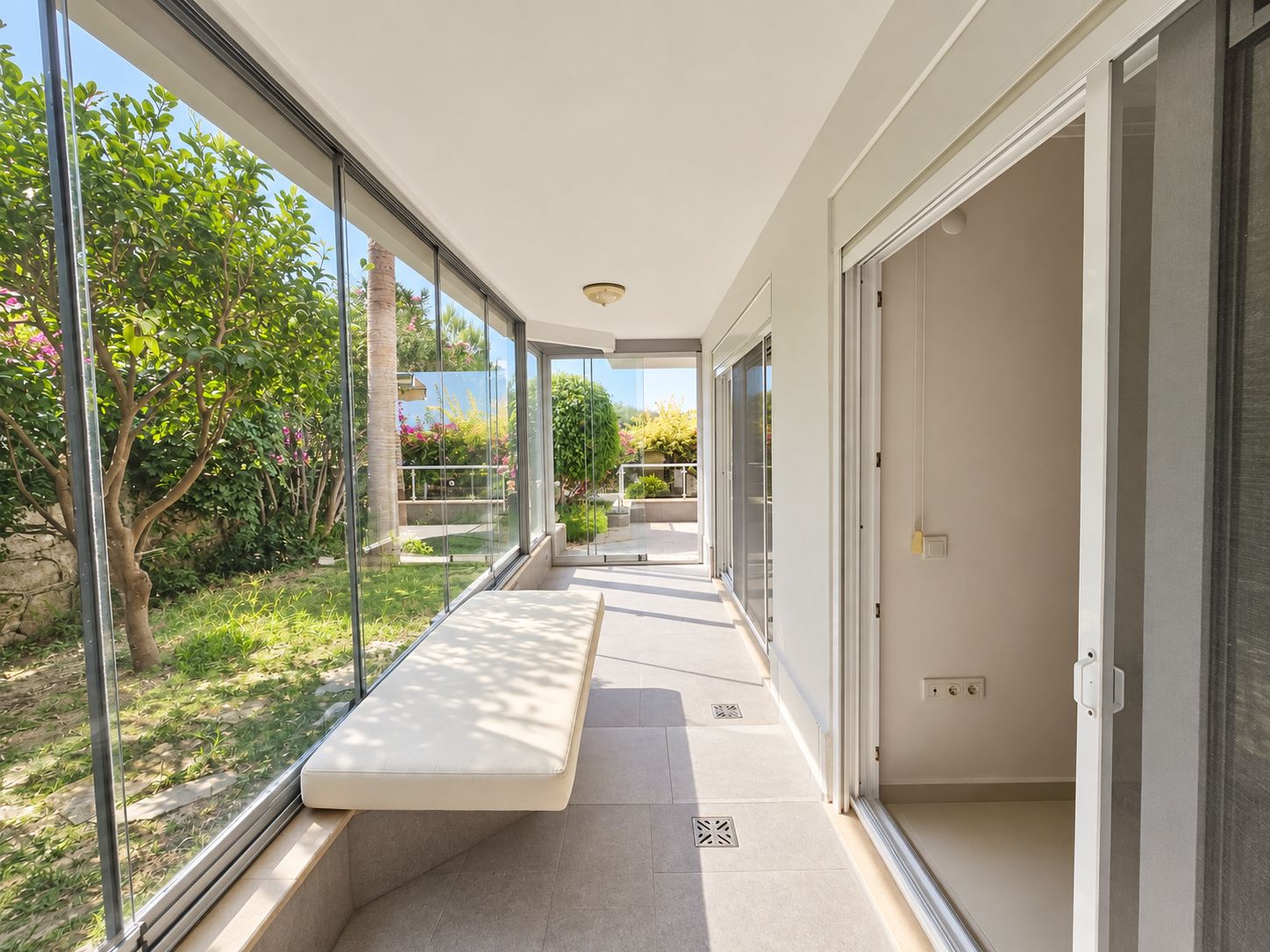 Long glazed terrace with a cushioned bench and trees beyond the windows in a flat in Konakli, Turkey