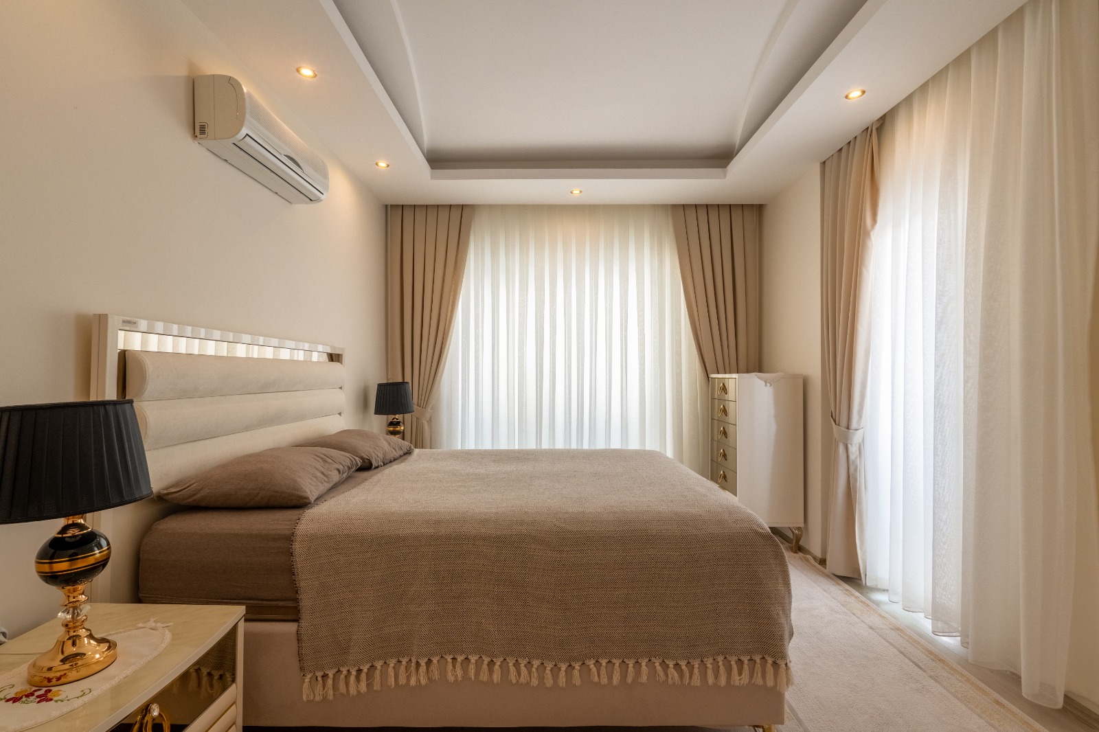 Двуспальная кровать с серо-бежевым покрывалом и белый комод в спальне квартиры